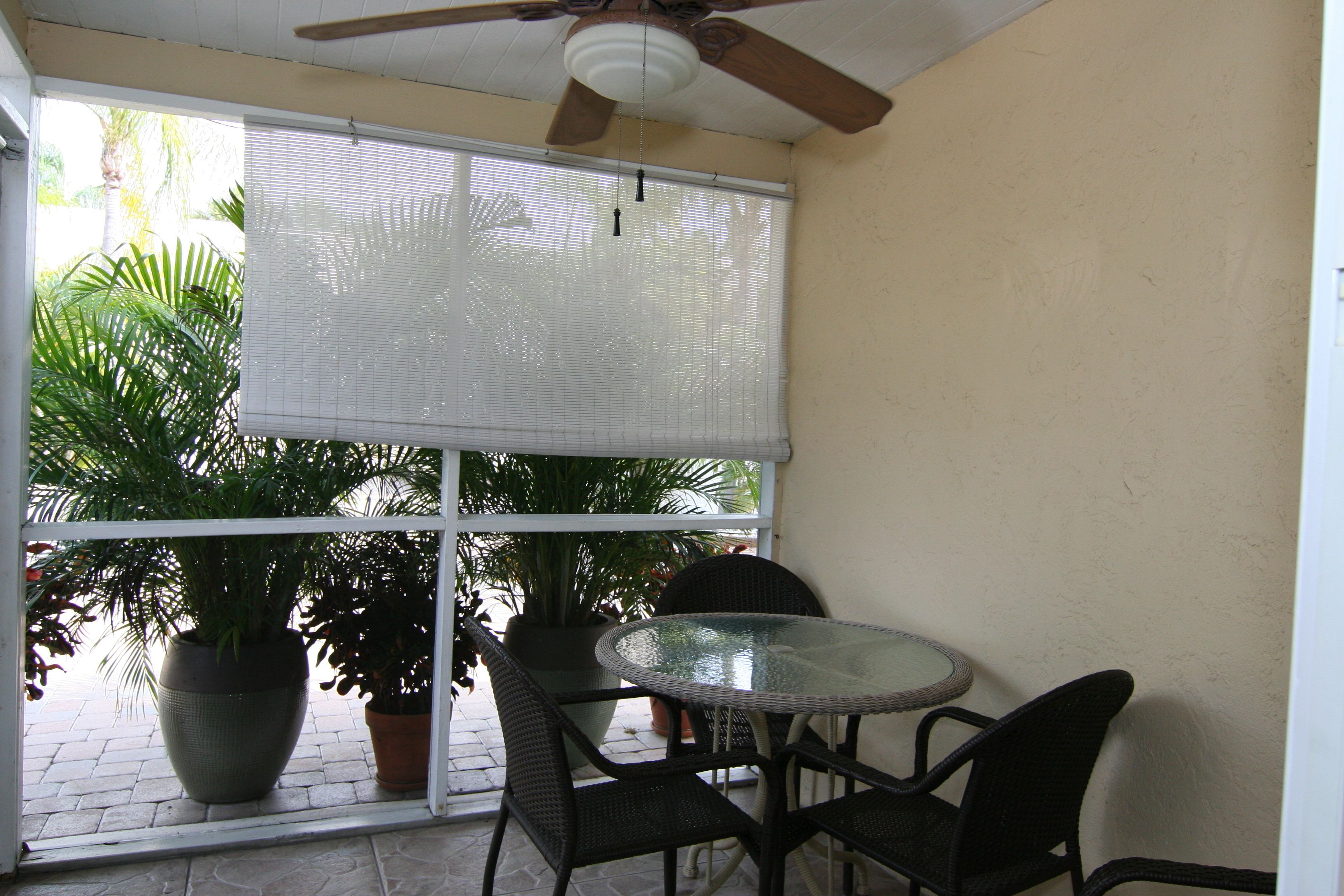 Une terrasse avec une table ronde en verre et quatre chaises, un ventilateur de plafond et de grandes plantes en pot.