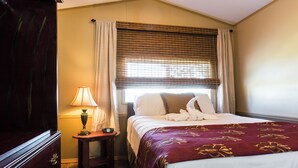 A hotel room with a bed, a nightstand with a lamp, a wooden dresser, and a window with curtains. - Red Shoes RV Park (Kinder)
