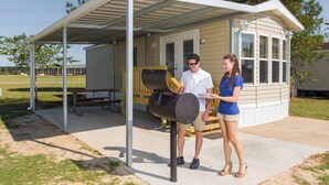 A covered outdoor area with a grill, a bench, and a small building with a door and windows. - Red Shoes RV Park (Kinder)