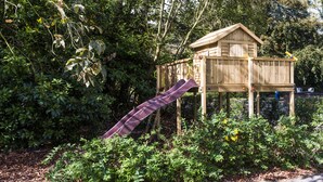 Une cabane en bois avec un toboggan, entourée d’un feuillage et de plantes luxuriants.