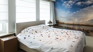 Une chambre avec un grand lit, une fenêtre avec des stores, une table de chevet, une lampe et une fresque représentant un paysage sur le mur.