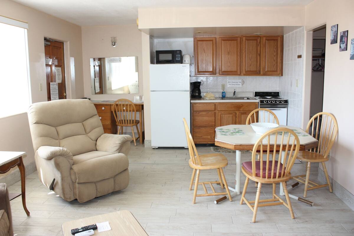 A compact living space with a sofa, dining table, and chairs, a kitchenette with a stove and microwave, and wooden cabinets.