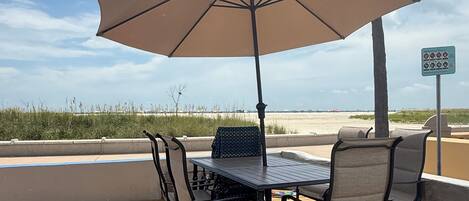 Une terrasse en bois avec une table et des chaises, protégée par un grand parasol, donnant sur une plage où l’on aperçoit un palmier et, au loin, un panneau.