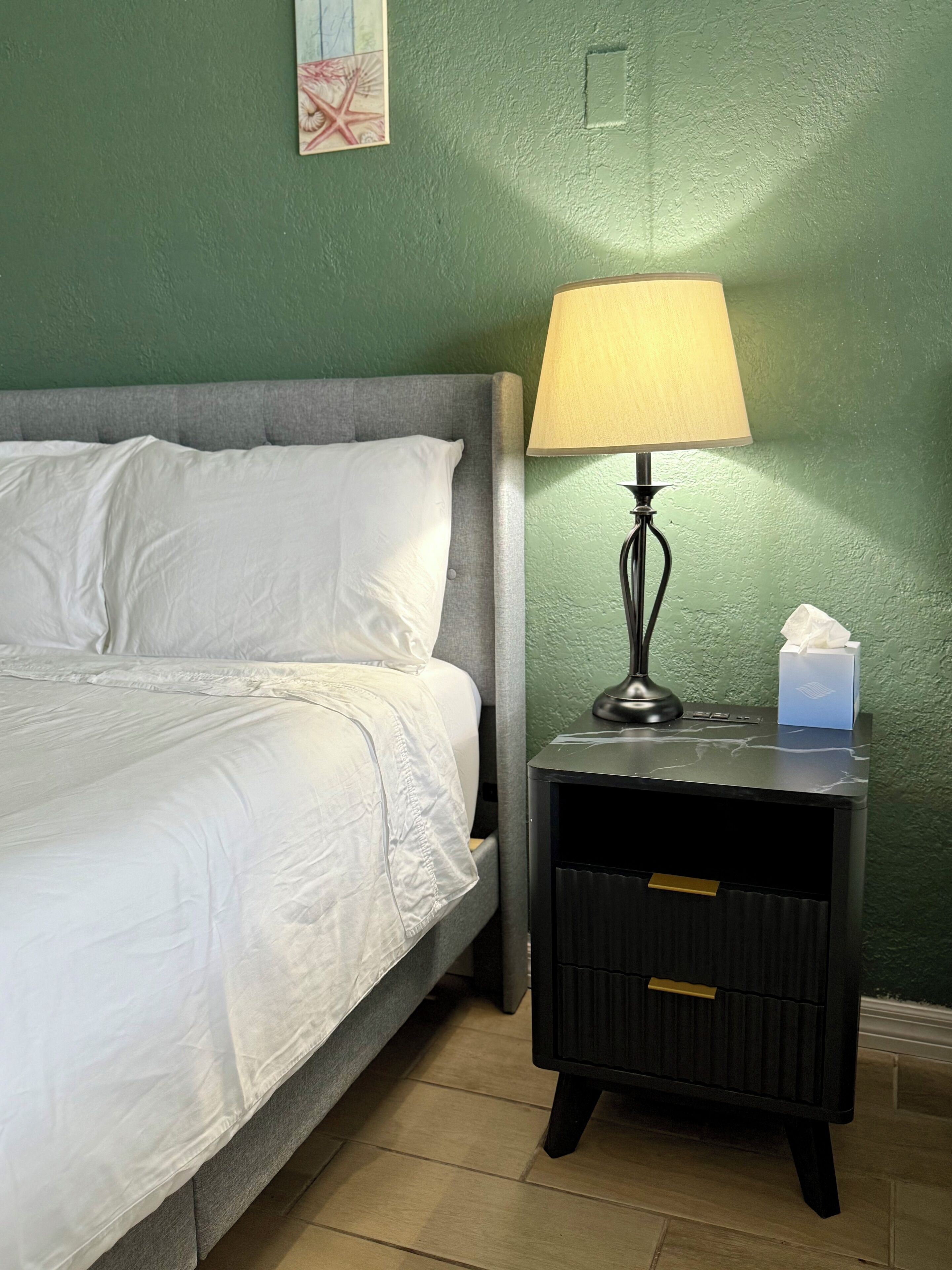 Un lit bien fait, avec du linge de lit blanc, une lampe de chevet, une boîte de mouchoirs et une petite commode.