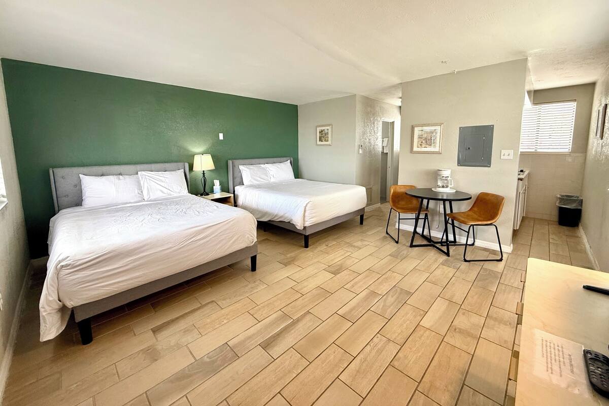 A hotel room with two beds, a small table with chairs, a coffee machine, and a trash bin.