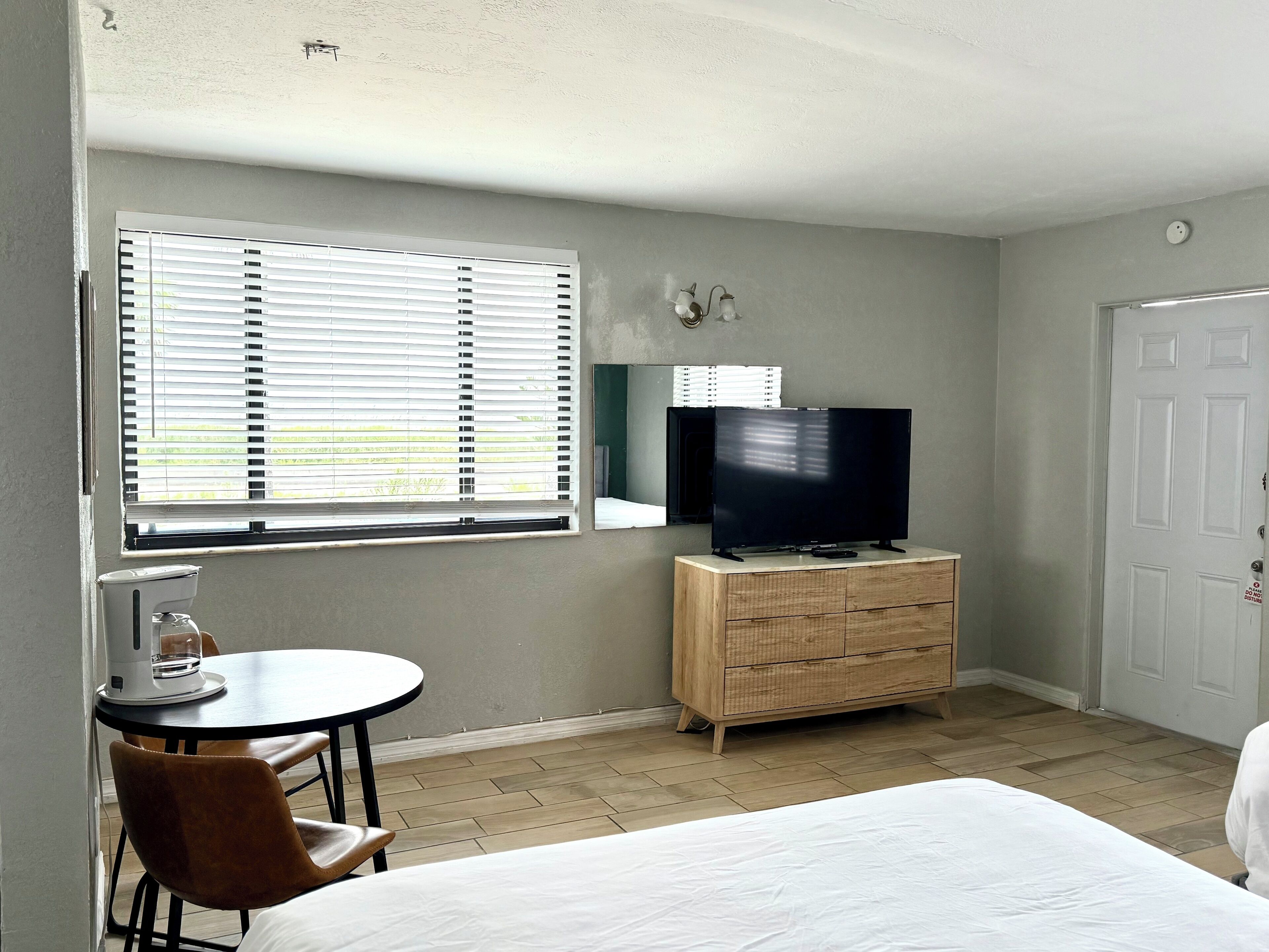 A room with a bed, a TV on a wooden dresser, a small table with a chair, and a window with blinds.