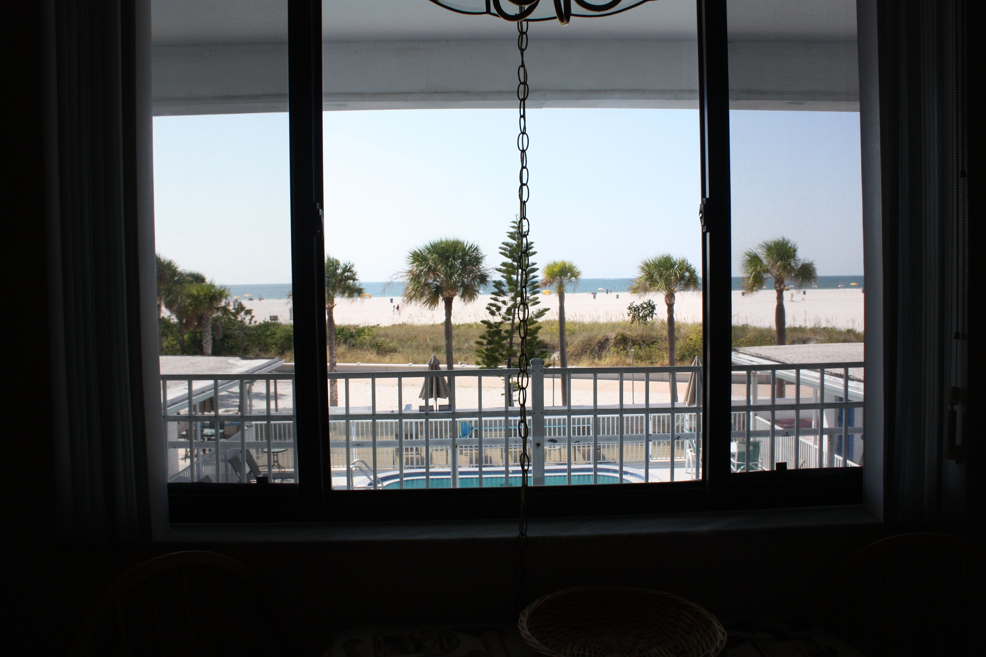 Vue depuis une chambre dotée d’une grande fenêtre donnant sur une plage, une piscine et des palmiers.