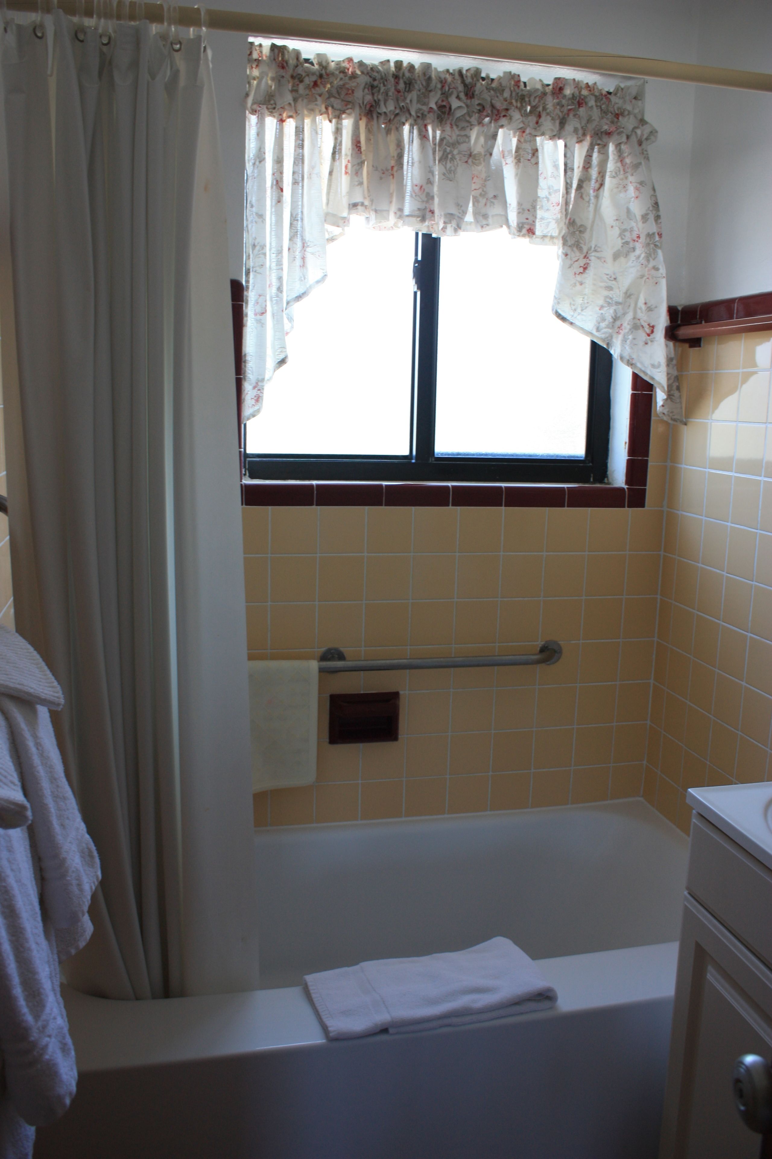 Un baño con bañera, una ventana con cortina floral, un perchero para toallas y una pared con azulejos.