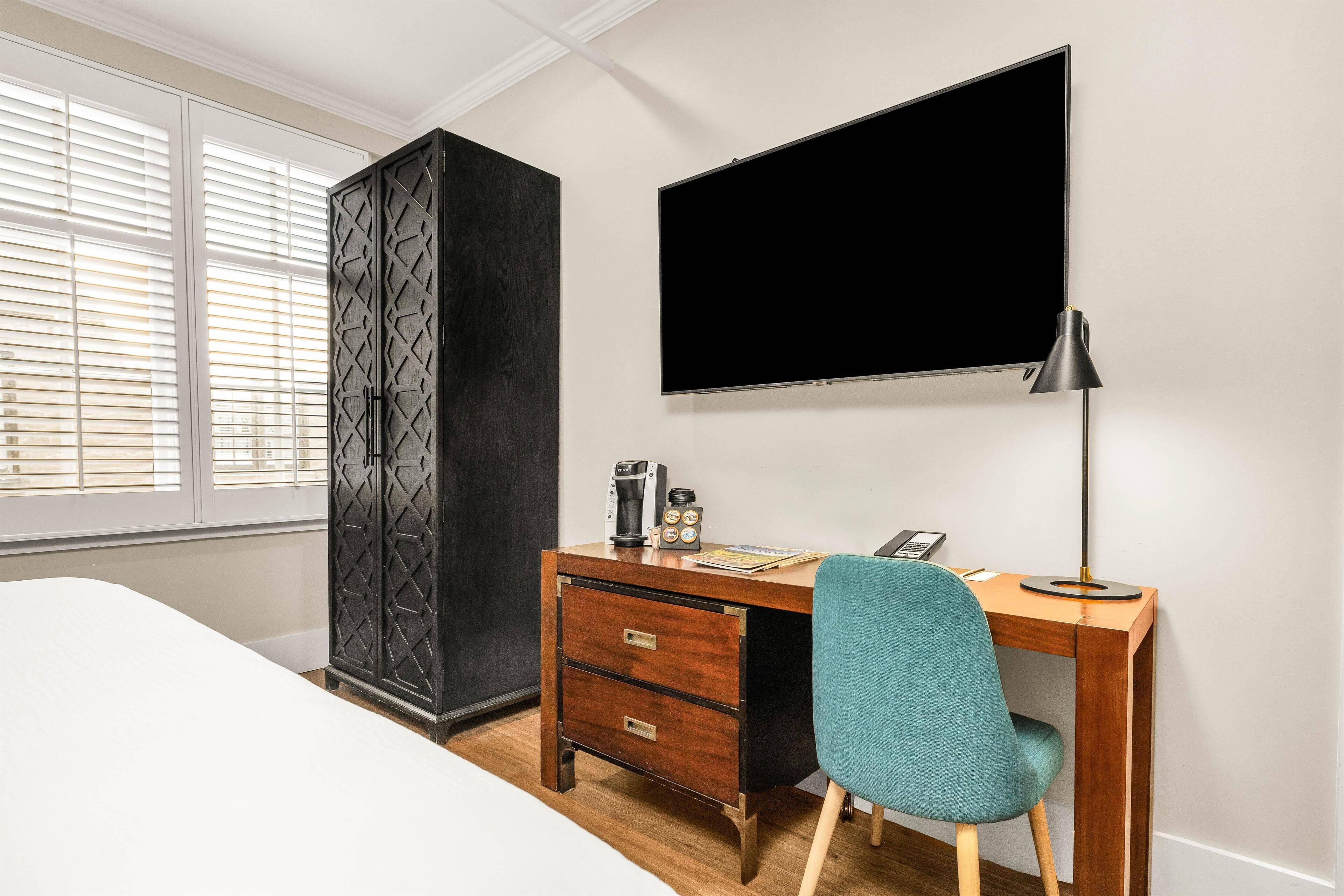 A hotel room with a bed, a desk with a chair, a TV, a wardrobe, and a window with blinds.