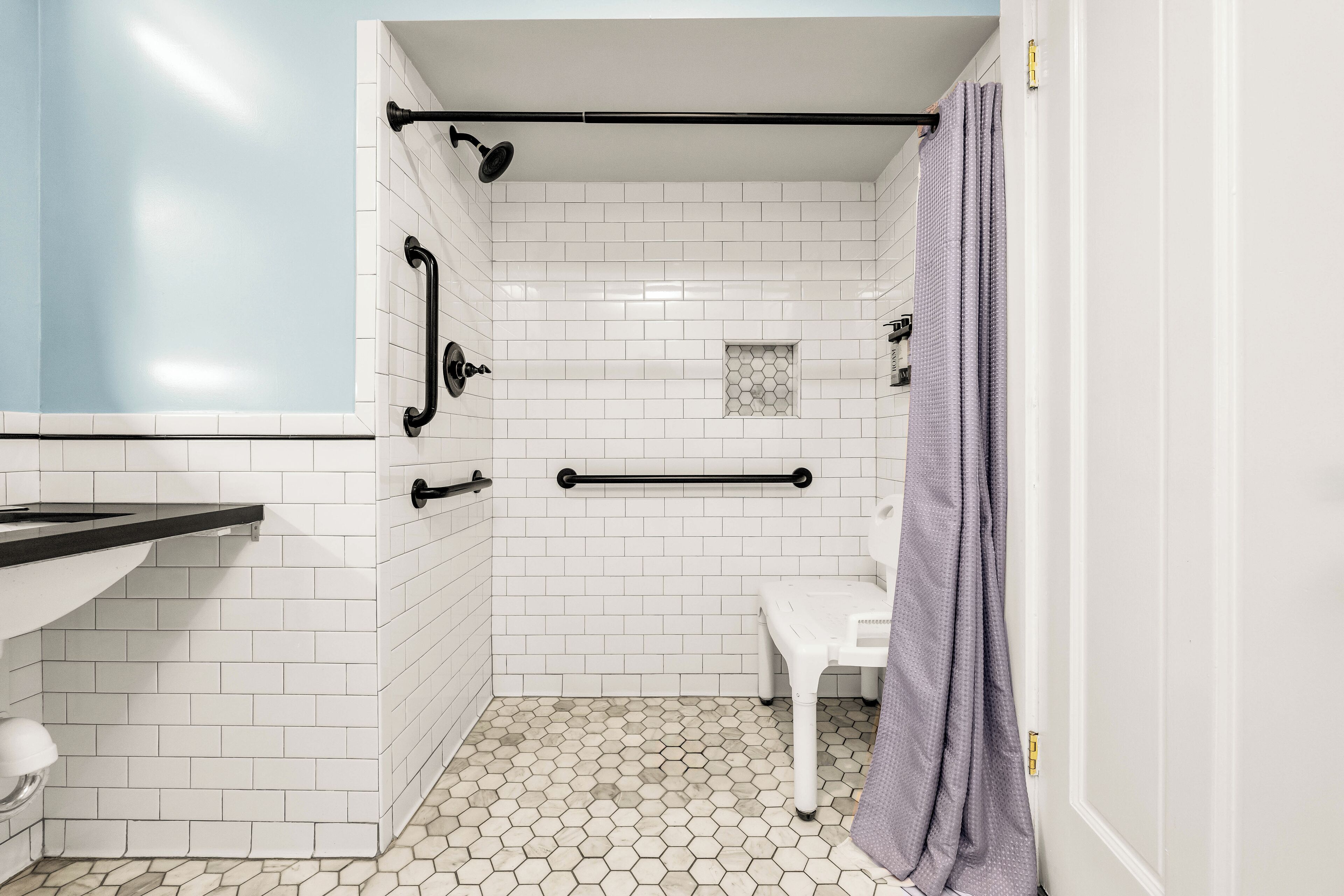 Une salle de bain avec des carreaux de métro blancs, un sol à motifs hexagonaux, un banc blanc et un rideau de douche violet.