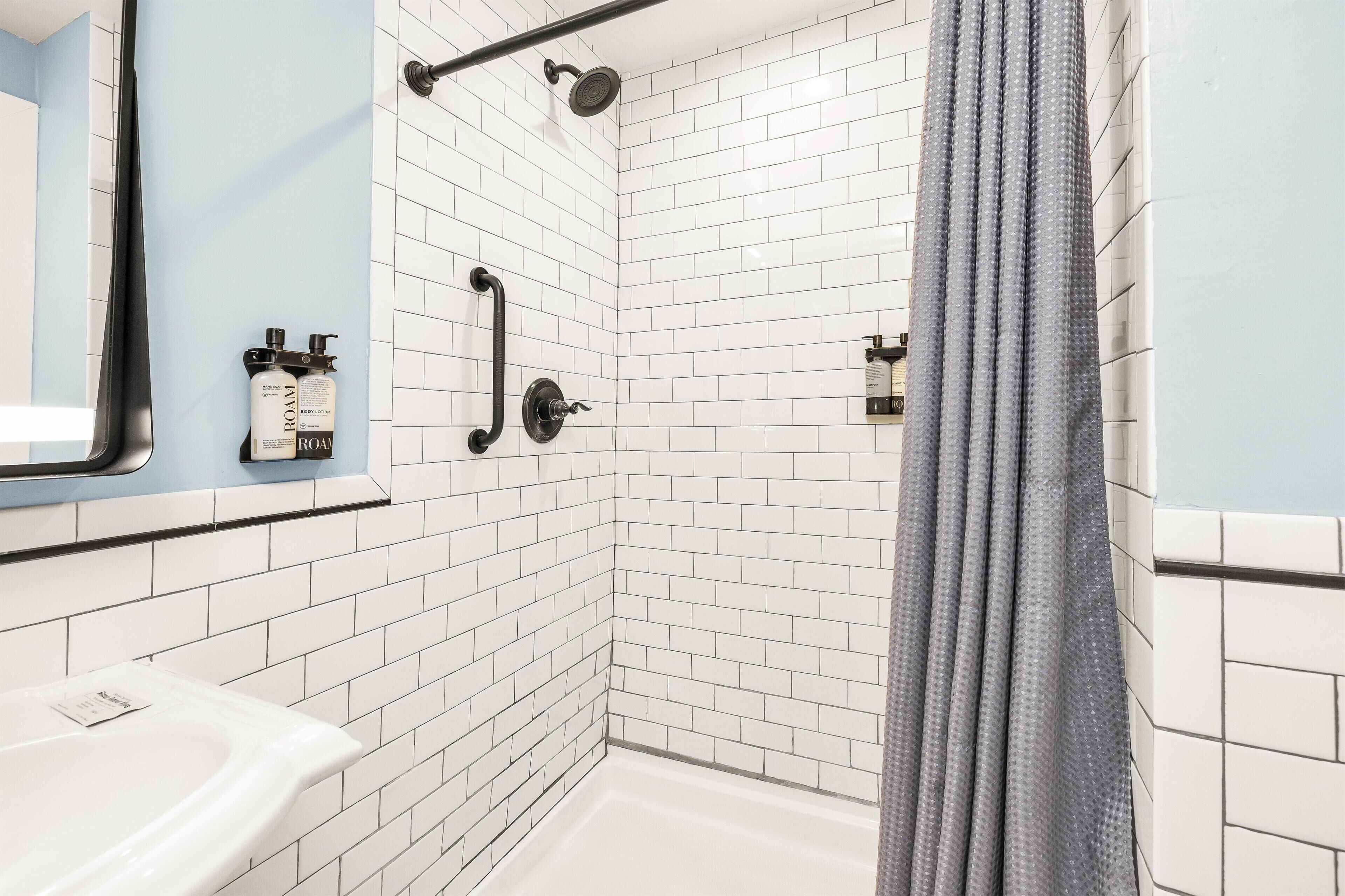Un área de ducha con paredes de azulejos blancos, un cabezal de ducha negro y una cortina de ducha gris.