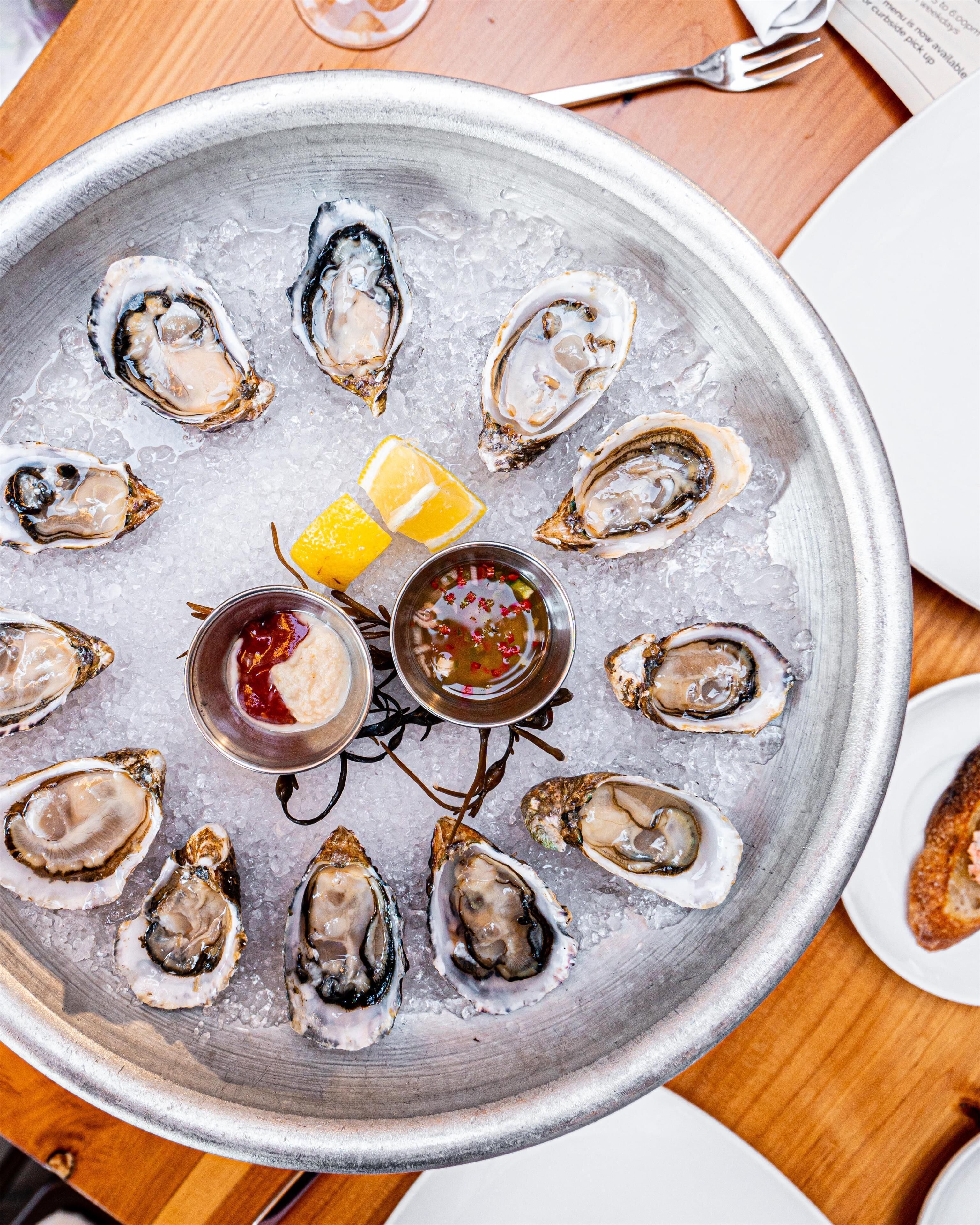 A tray of oysters on ice with lemon wedges and two small bowls of sauce.