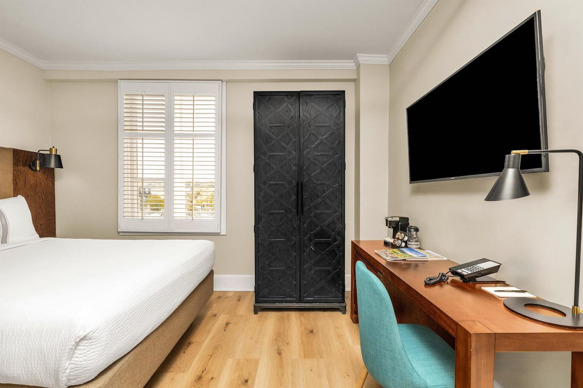 A hotel room with a bed, desk, chair, TV, wardrobe, and window.