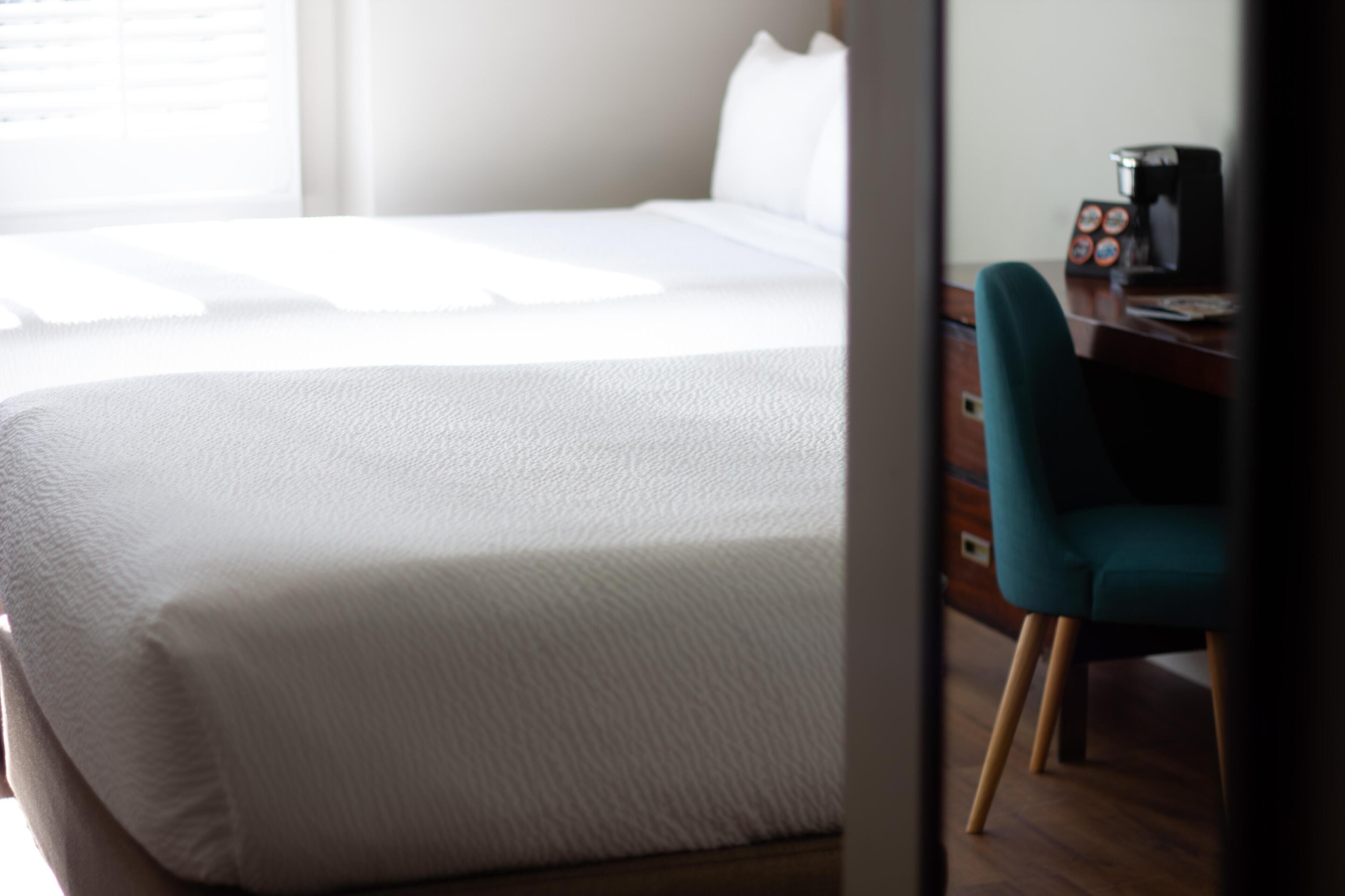 A neatly made bed with white bedding, a teal chair, and a wooden desk with a coffee machine.