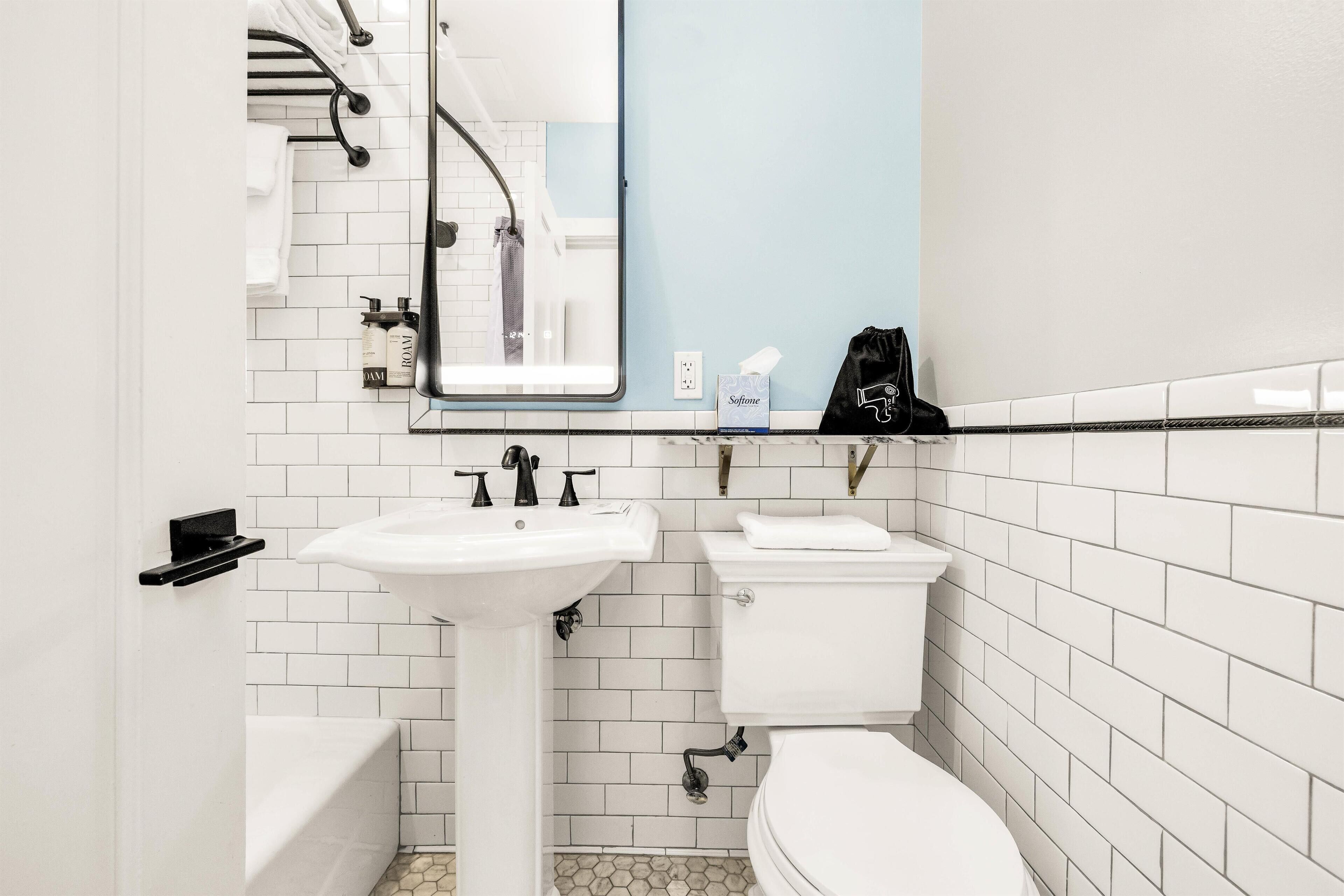 Une salle de bain avec un lavabo, des toilettes et une baignoire, tous de couleur blanche. On trouve un porte-serviettes au-dessus du lavabo et un miroir sur le mur.