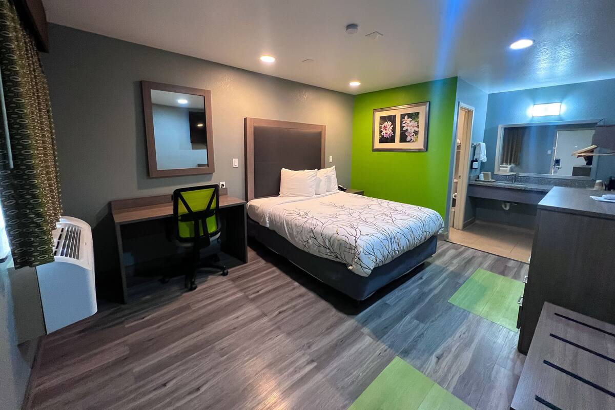 Quarto de hotel com cama, secretária, cadeira, espelho e área de banheiro.