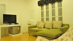 A room with a green sofa, a television on a stand, and a window with patterned curtains. - Boutique Guesthouse by Clearhouse (Kamala)