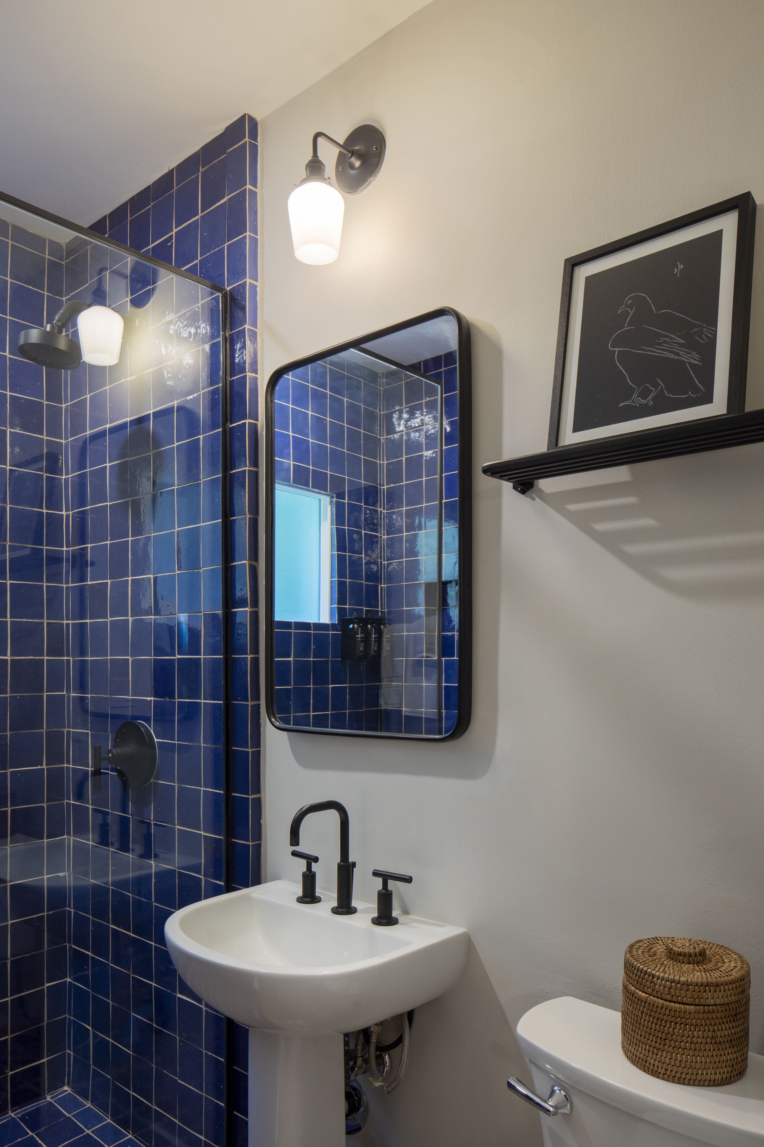 Ein Badezimmer mit einem weißen Waschbecken, einem Spiegel, einem gerahmten Bild und einer Duschkabine mit blauen Fliesen.
