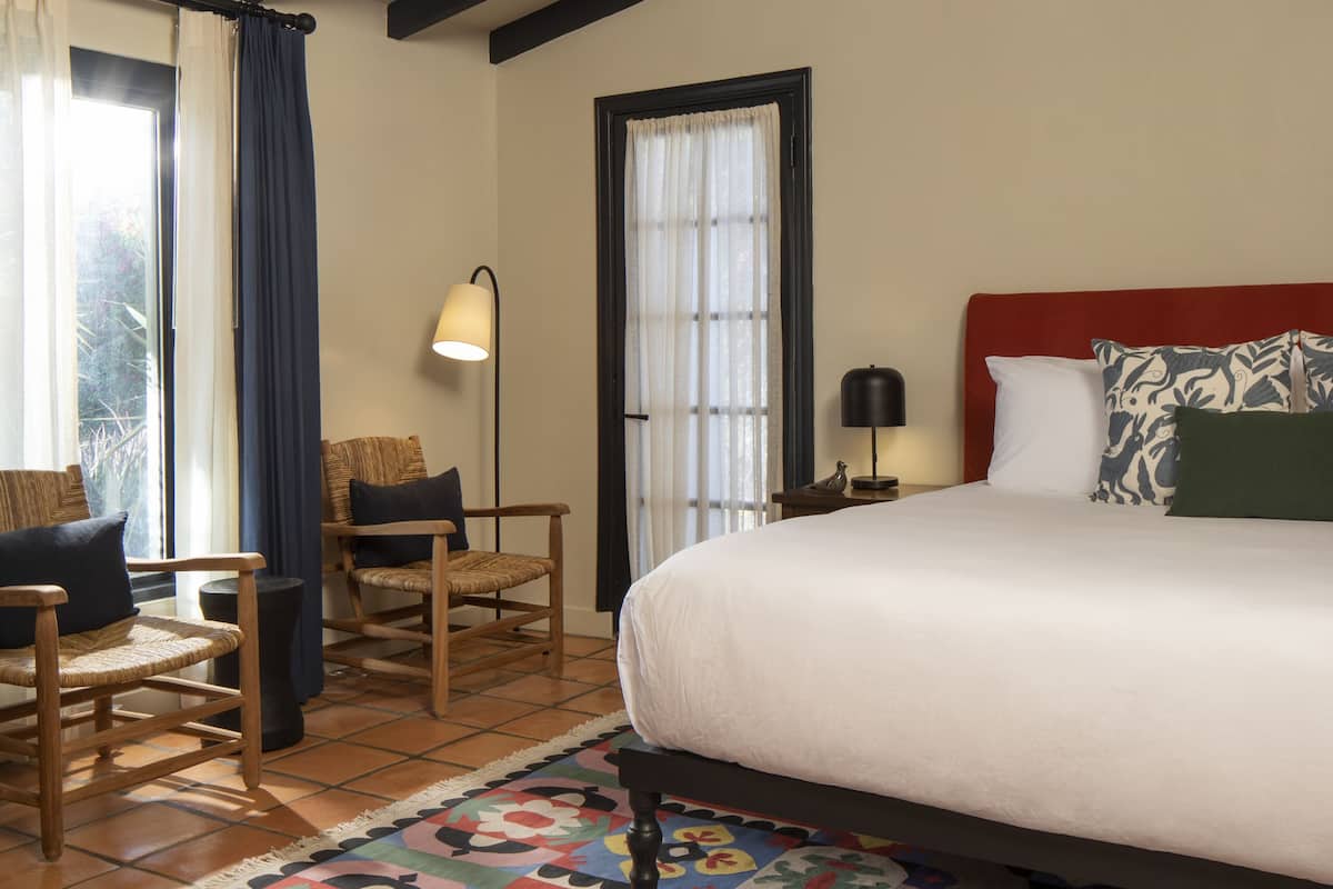 A bedroom with a large bed, a wooden chair, a floor lamp, and a patterned rug.