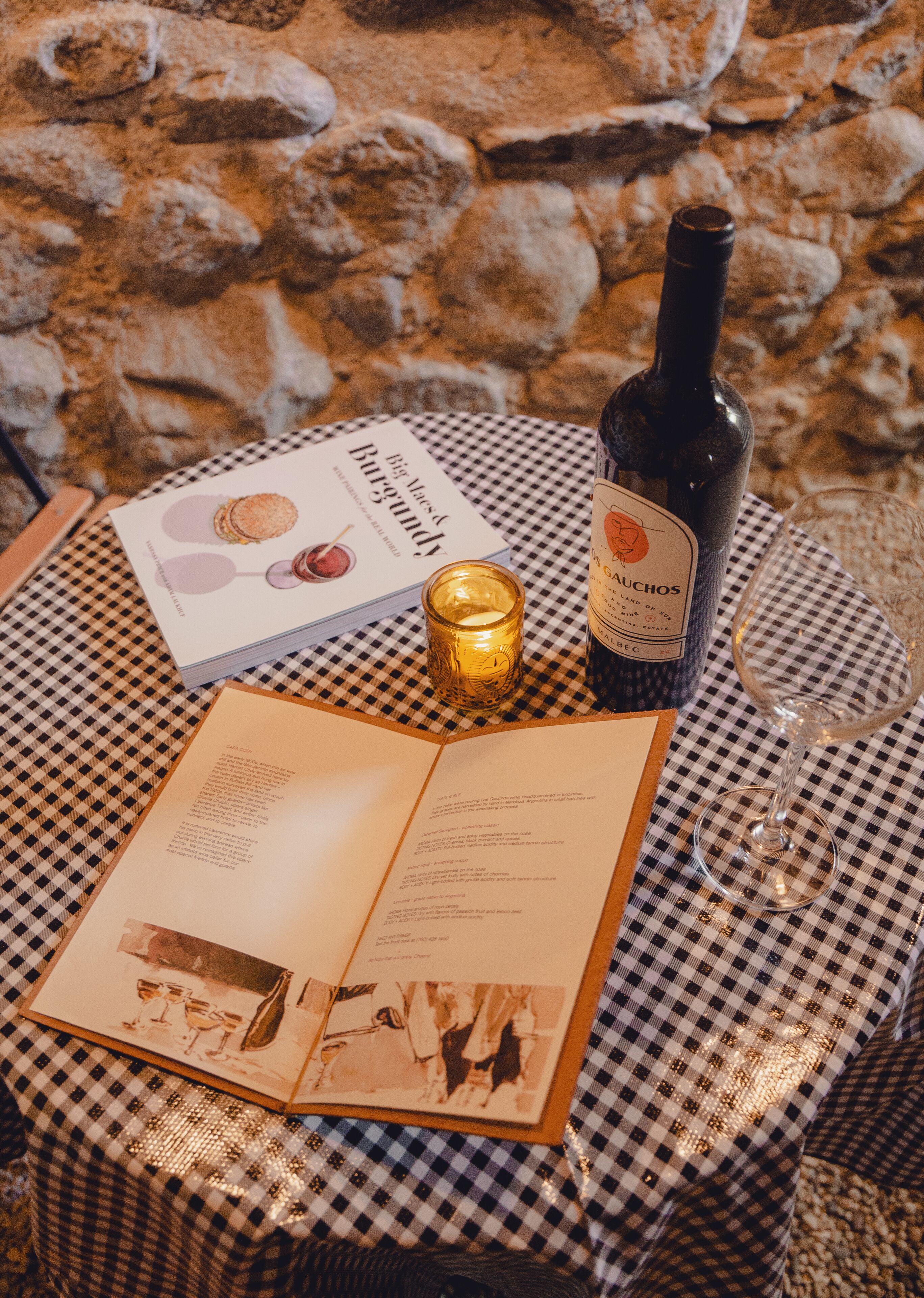 A table with a book, a bottle of wine, a candle, and a glass on a checkered tablecloth.