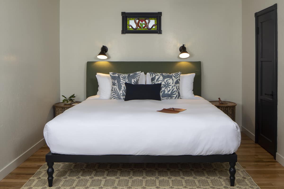 A neatly made bed with white bedding and patterned pillows, a green headboard, and two bedside tables with lamps.