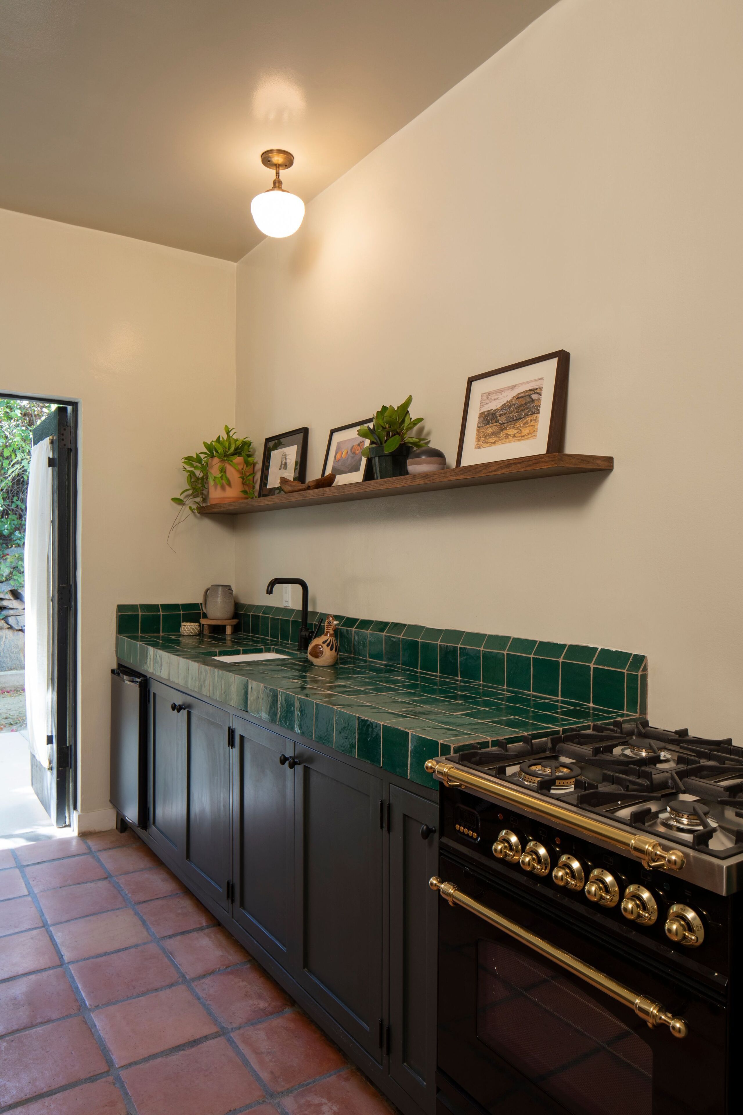 A kitchen with a green tiled backsplash, a black stove, and a wooden shelf above the sink.
