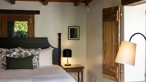 Une chambre avec un sol en pierre, un lit doté d’une tête de lit sombre, une table de chevet avec une lampe et une fenêtre avec un volet en bois.