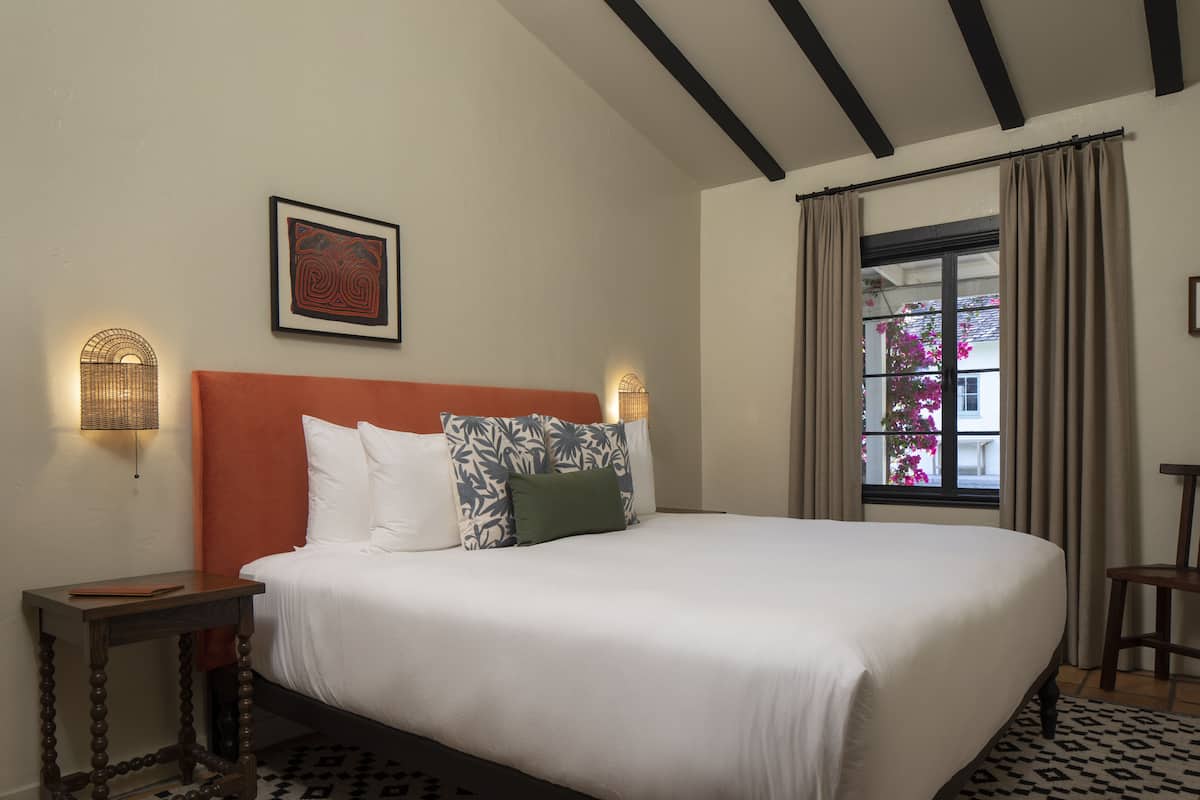 A neatly made bed with white linens and patterned pillows, a small bedside table with a book, and a framed artwork on the wall.