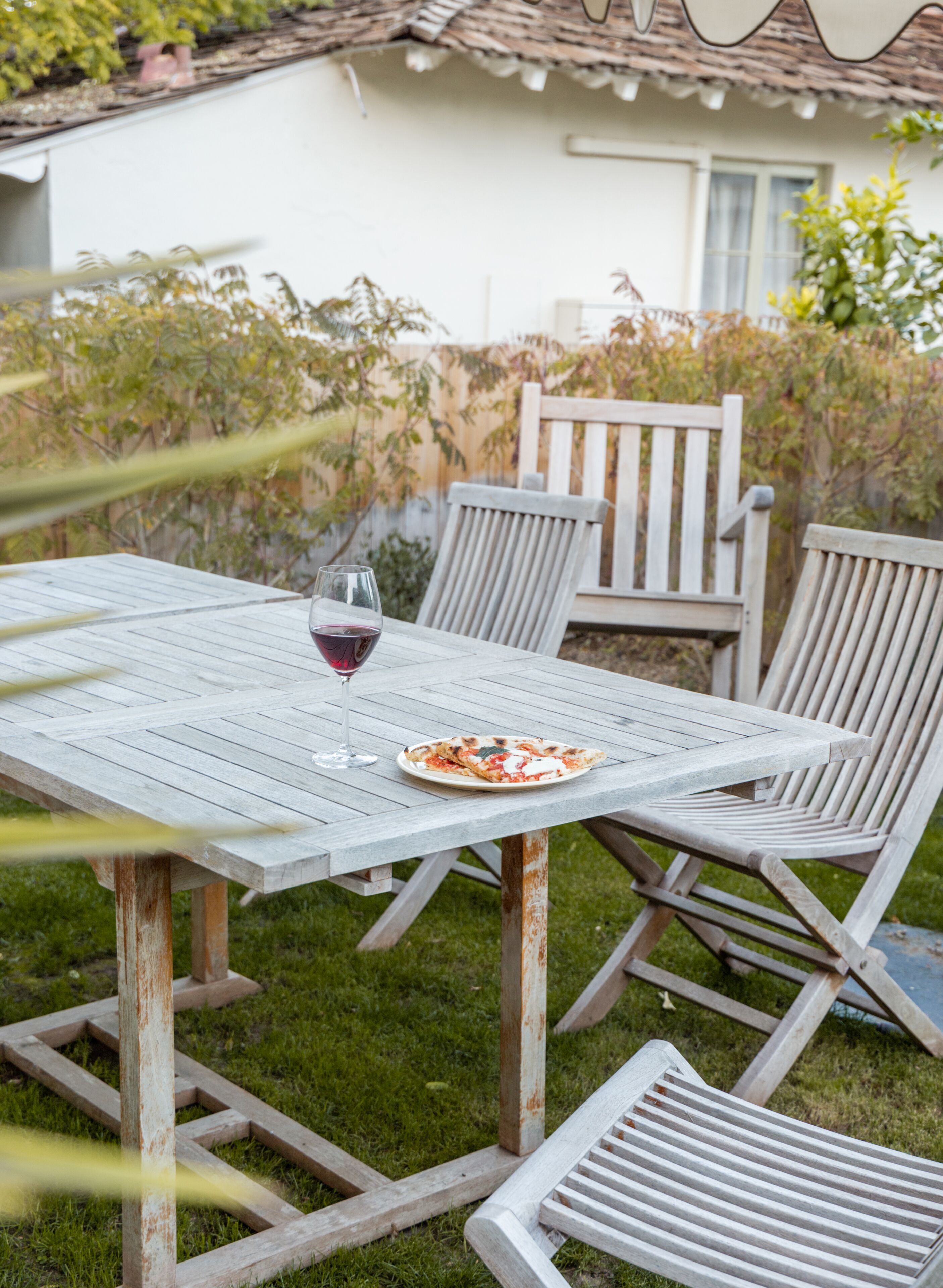 An outdoor wooden table with chairs, a glass of red wine, and a plate of food.