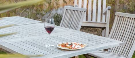 An outdoor wooden table with chairs, a glass of red wine, and a plate of food.