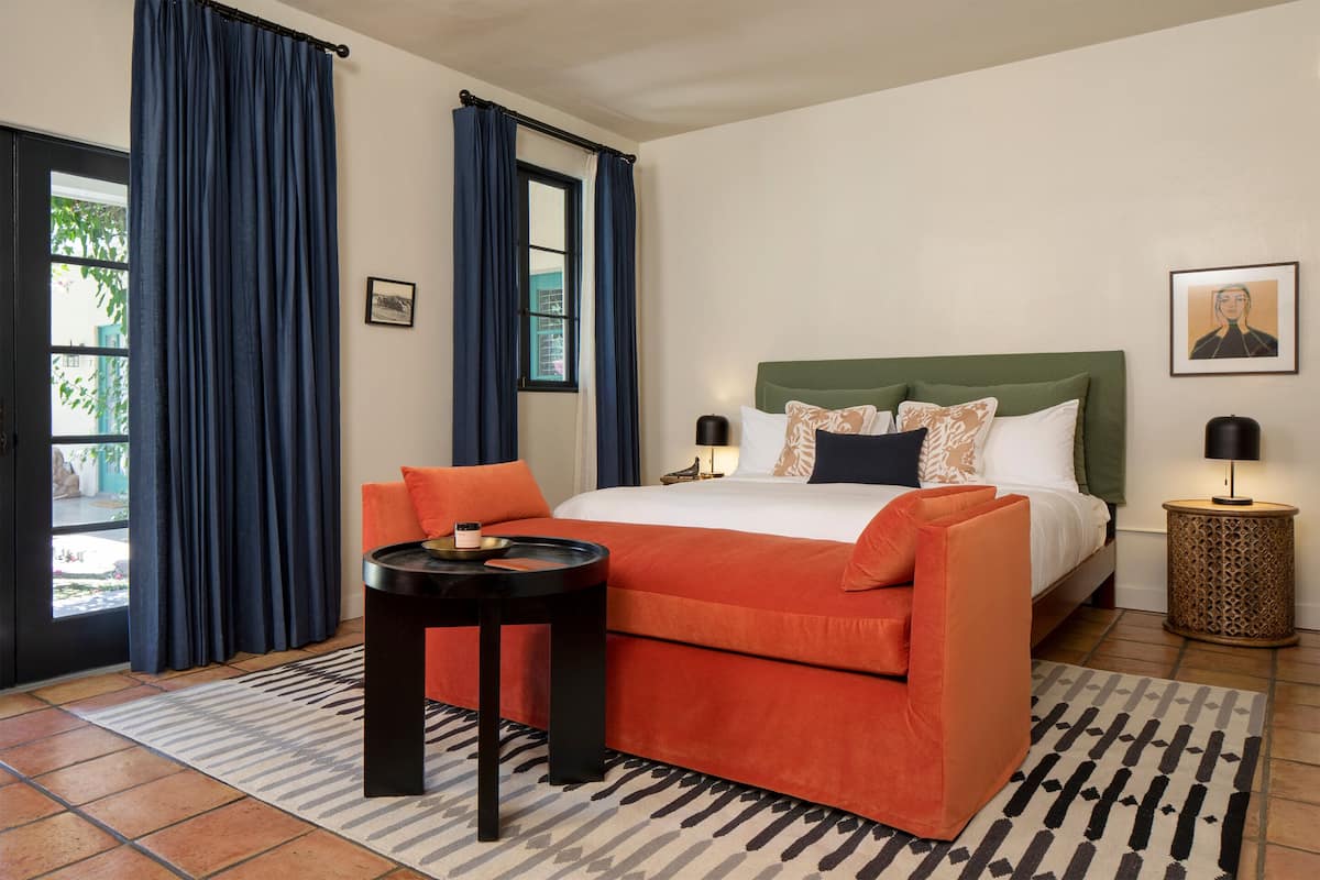 A hotel room with a large bed, orange and white bedding, a small round table, a patterned rug, and a view of the outdoors through a window with blue curtains.