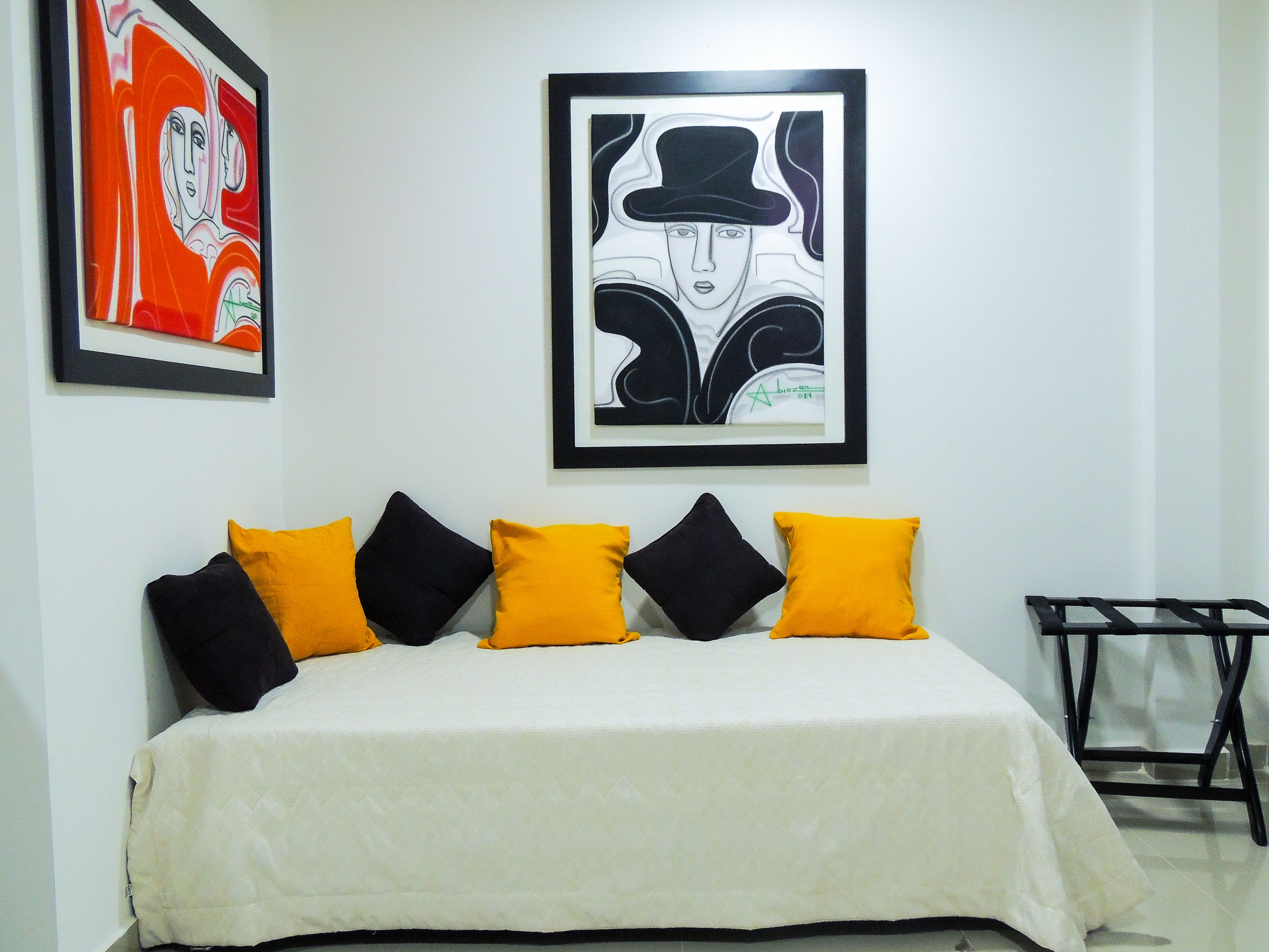 Una cama con cubierta blanca, almohadas negras y amarillas, y dos cuadros enmarcados en la pared.