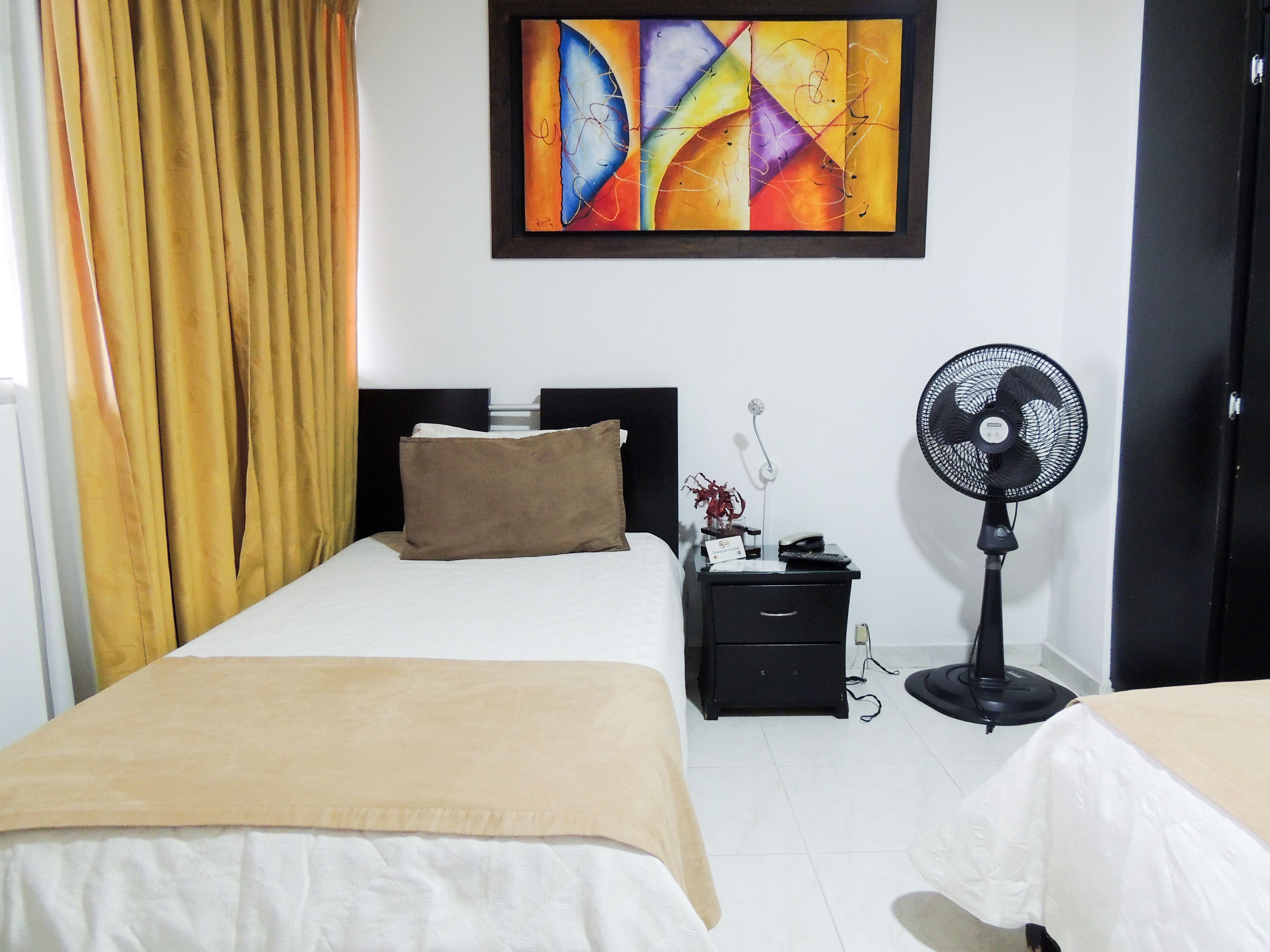 Habitación de hotel con una cama, una mesita de noche con teléfono y ventilador, un cuadro en la pared y una ventana con cortinas.