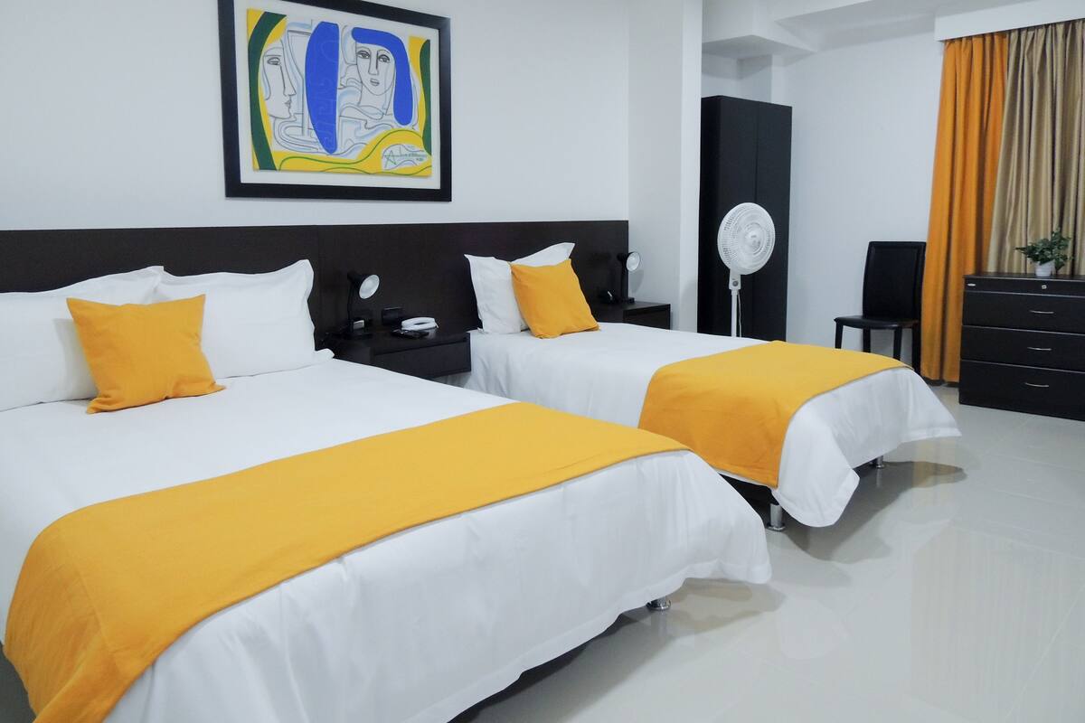 Ein Hotelzimmer mit zwei Betten, einem Nachttisch, einem Schreibtisch, einem Stuhl, einem Ventilator und einem Bild an der Wand.