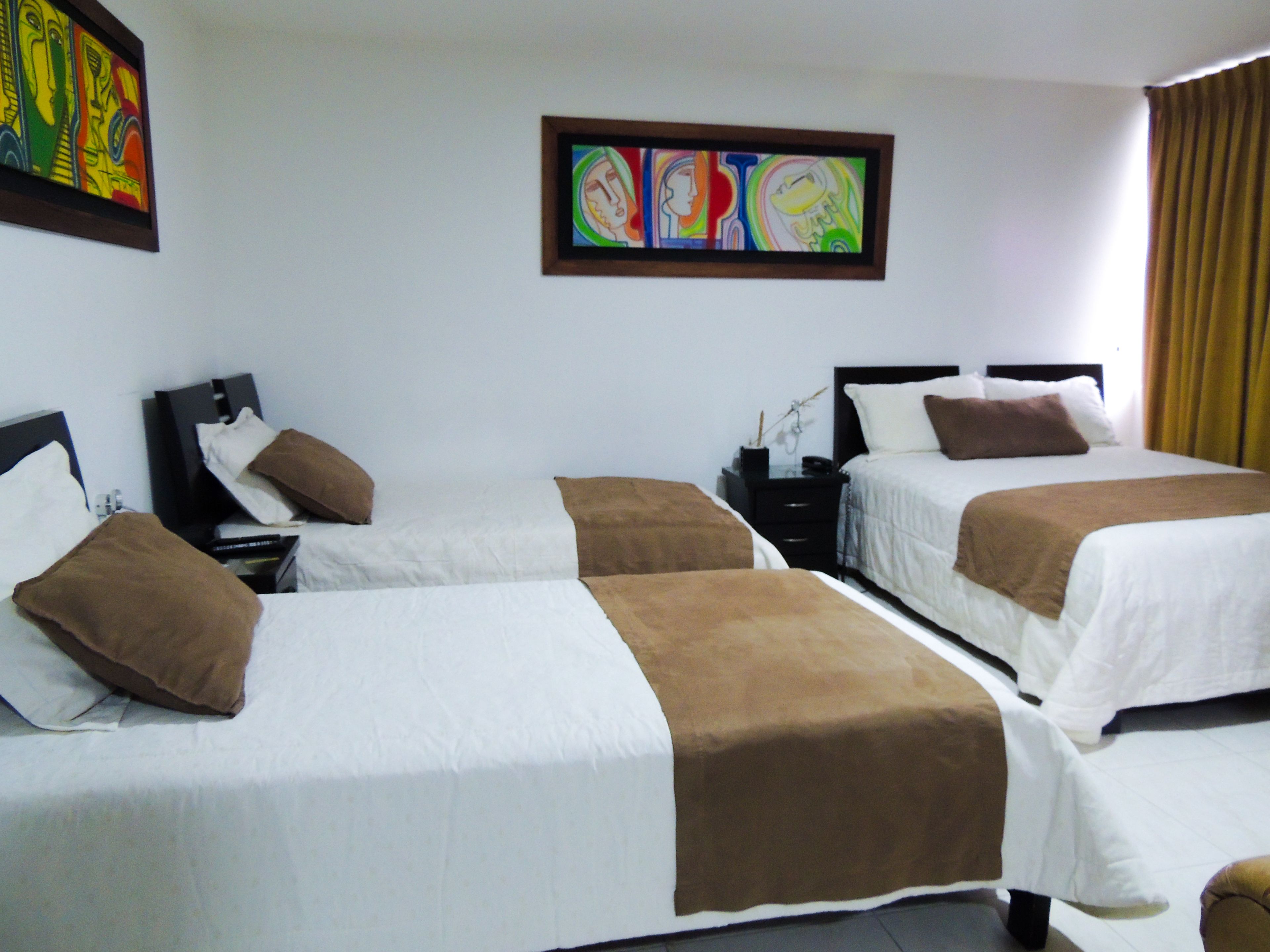A hotel room with two beds, each with white bedding and brown throw pillows. There is a framed abstract painting above the beds, and a nightstand with a lamp and a phone on it.