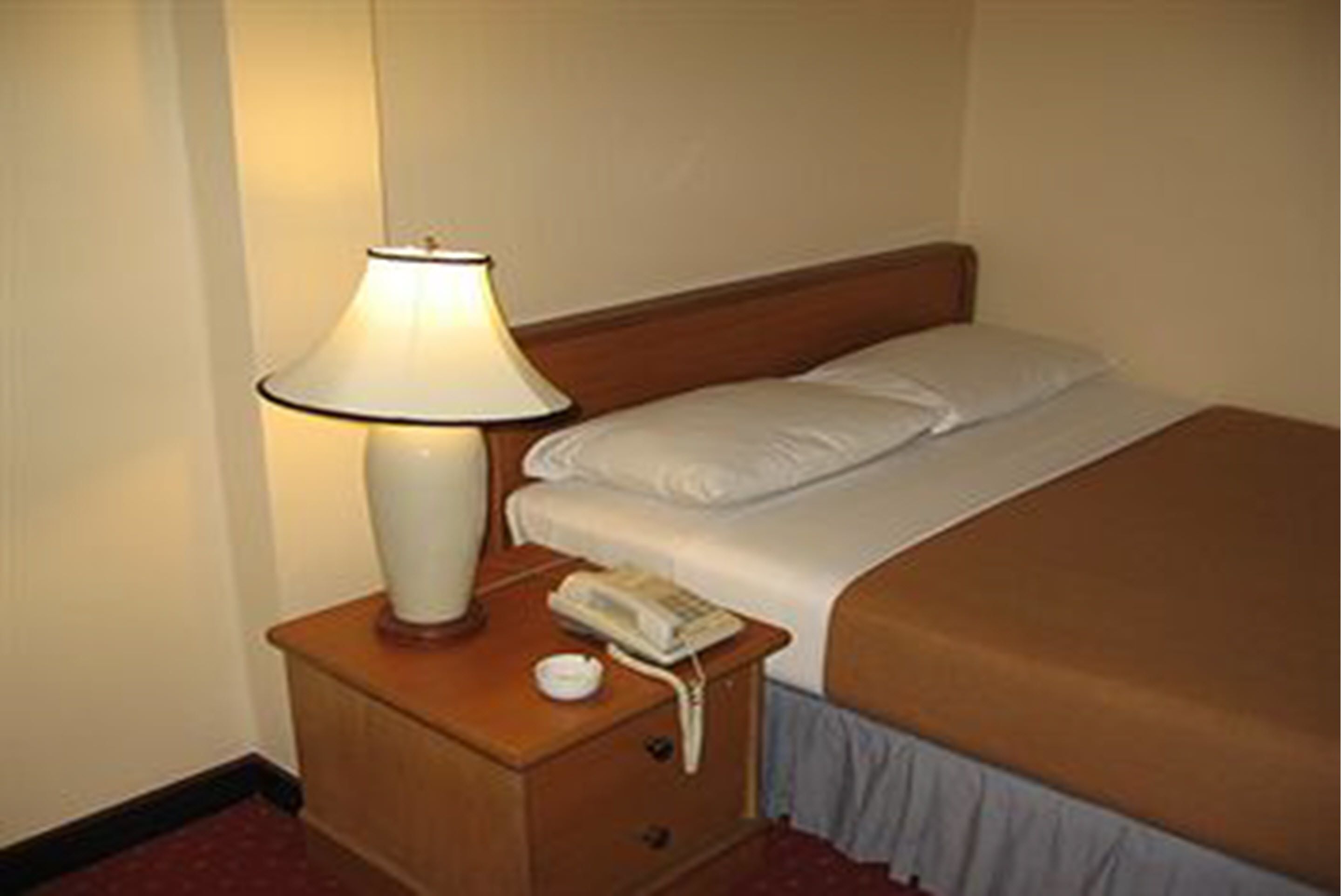 A hotel room with a bed, a bedside lamp, a telephone, and a small table with a cup.