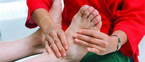 A person in a red top and green pants performing a foot massage on another person.