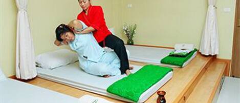 A room with two beds, a small table with a bowl of flowers, and a person sitting on one bed.
