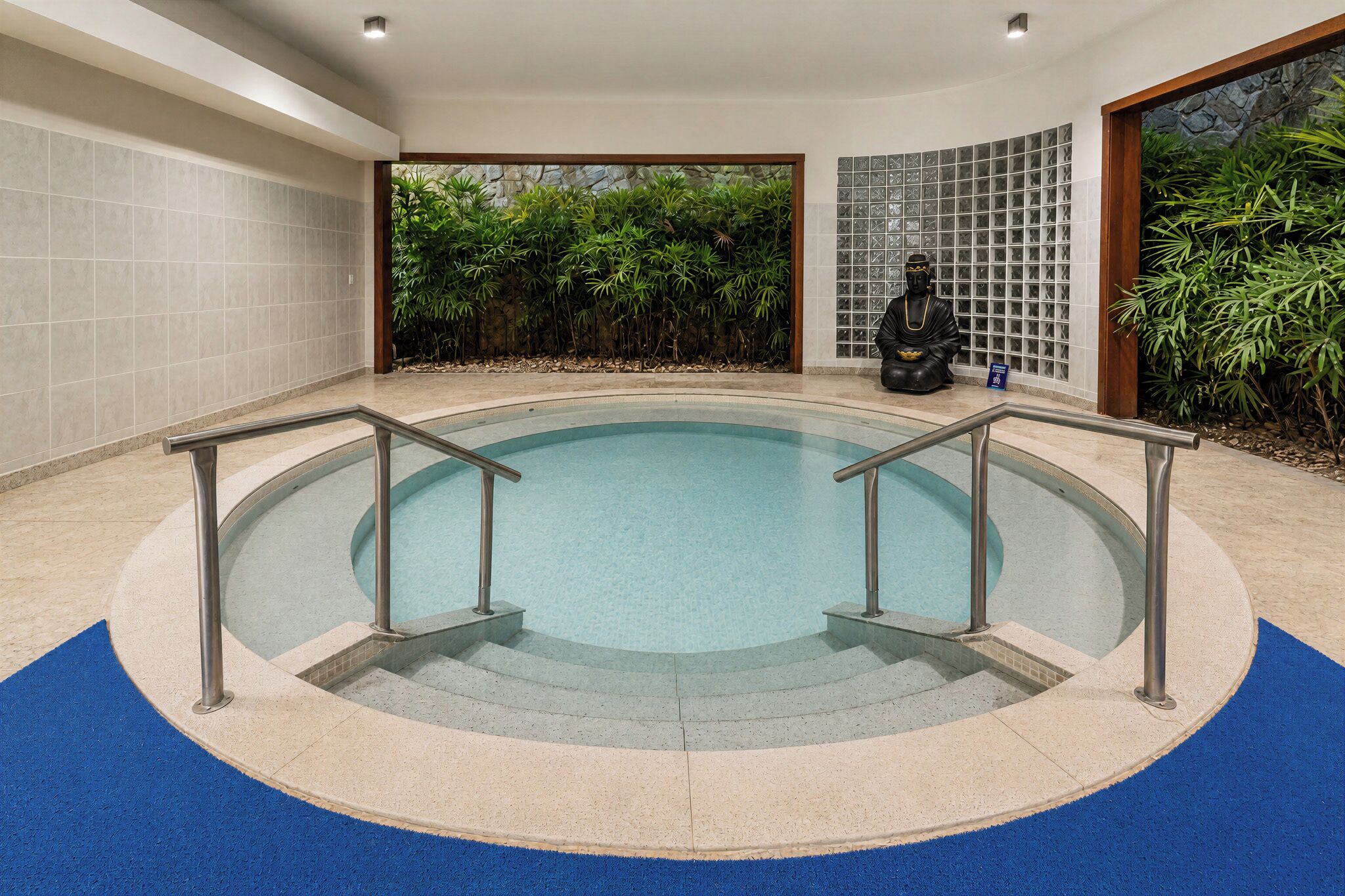 Ein runder, überdachter Pool mit Metallgeländern, umgeben von einem blauen Teppichbereich, mit einer Steinwand und Grünpflanzen im Hintergrund.