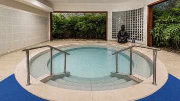 Ein runder, überdachter Pool mit Metallgeländern, umgeben von einem blauen Teppichbereich, mit einer Steinwand und Grünpflanzen im Hintergrund.