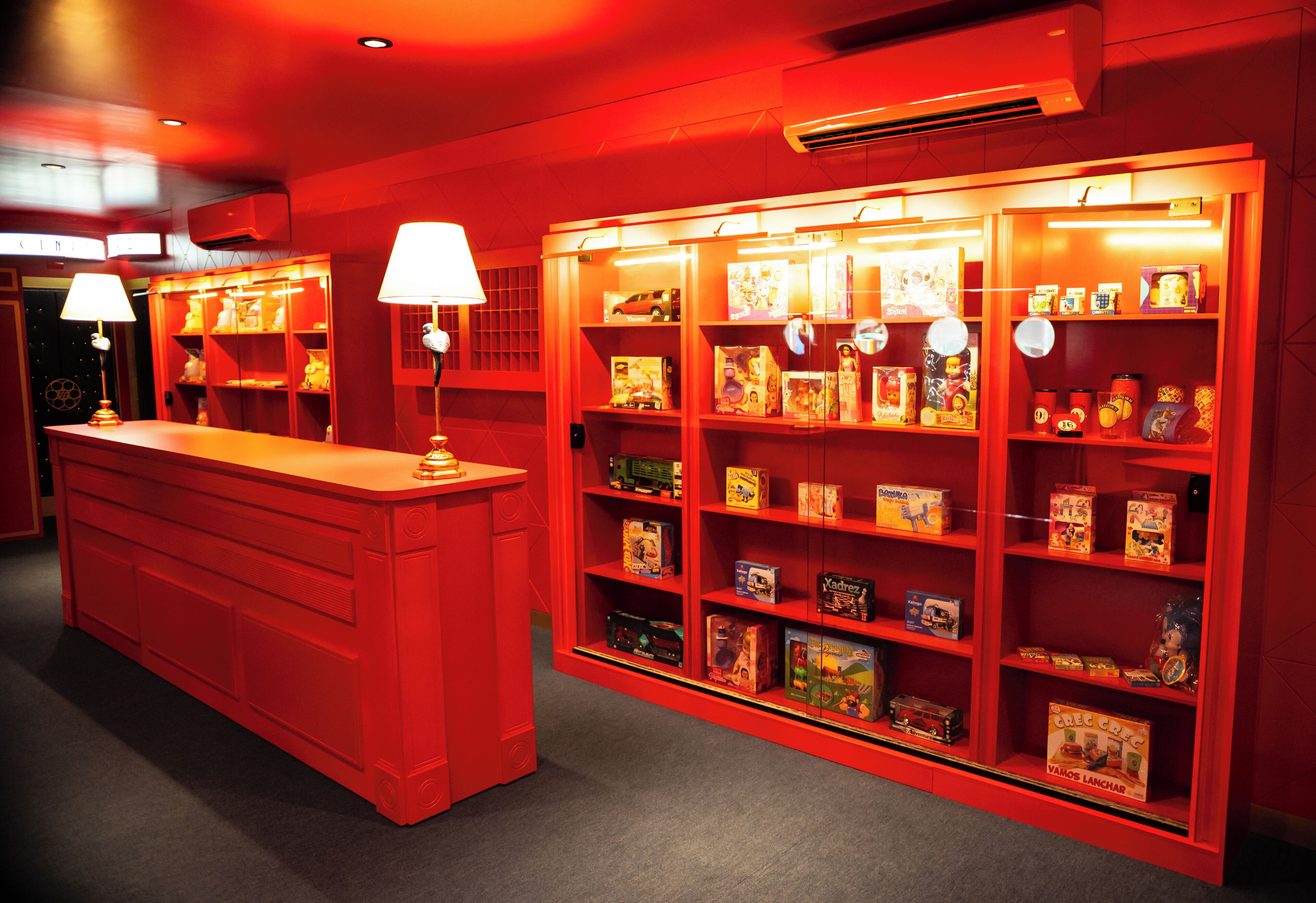 A red-themed room with a central desk, shelves with toys, and two lamps.