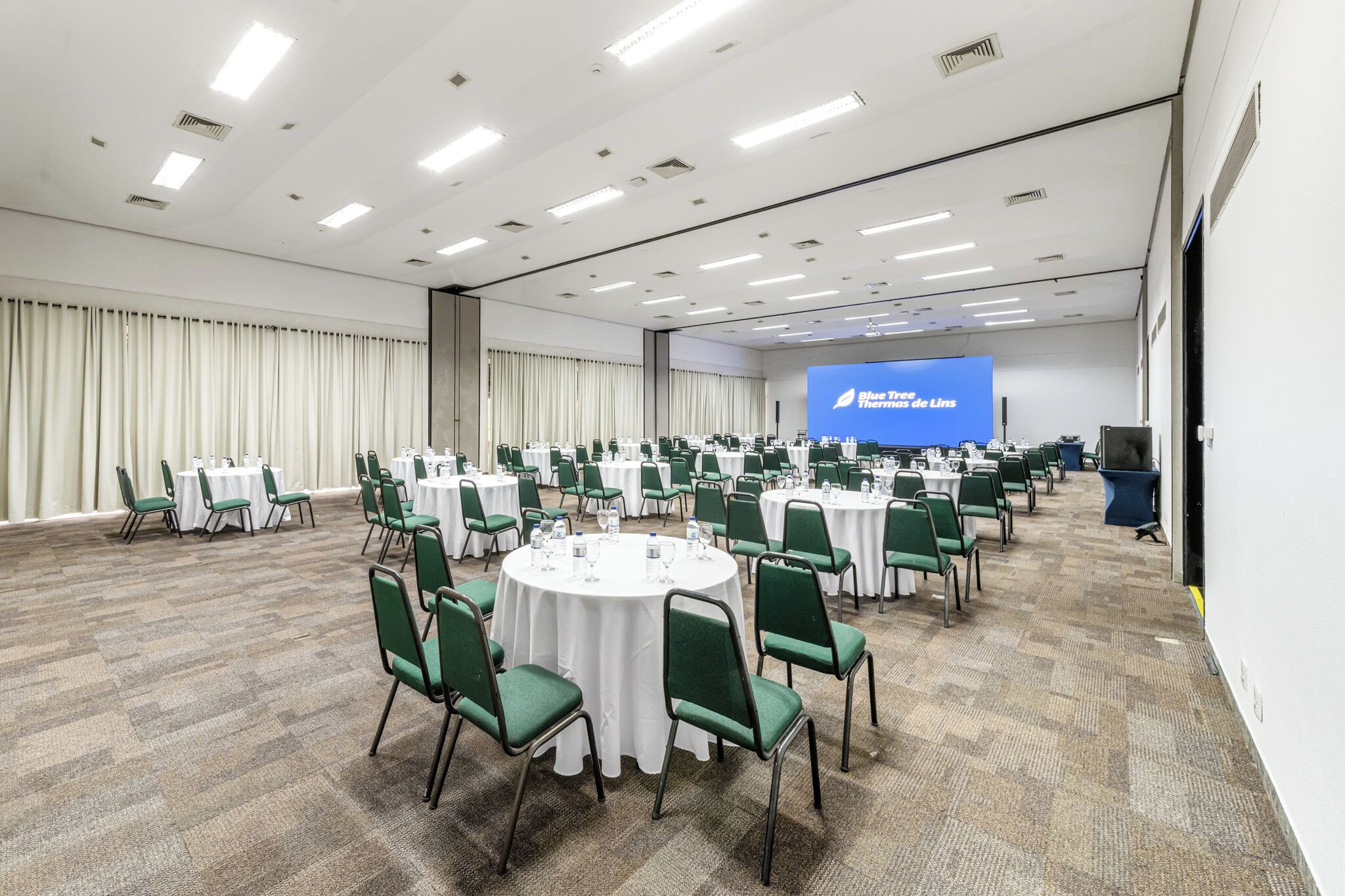 A large conference room with round tables, green chairs, and a projection screen displaying 'Blue Tree de Inns.'