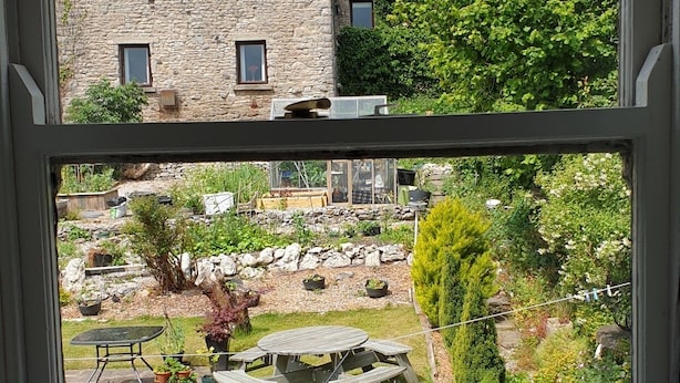 A stone building with four windows, a garden with a wooden table and chairs, and various plants.