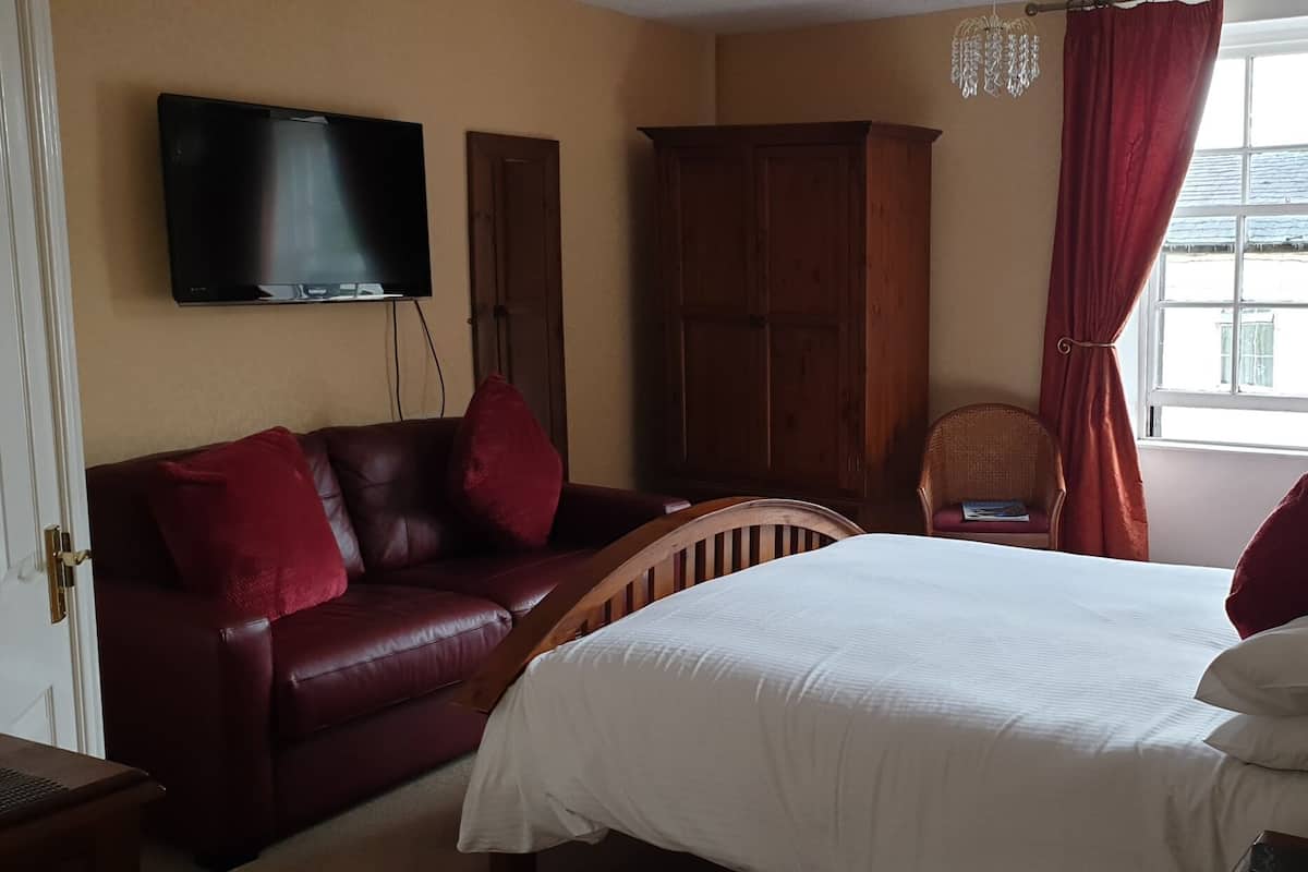 Habitación de hotel con cama, sofá de cuero rojo, armario de madera, televisión montada en la pared y ventana con cortinas rojas.