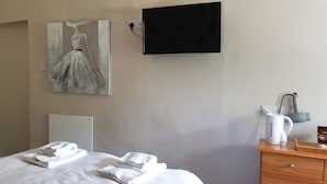 A hotel room with a bed, a painting of a dress on the wall, a TV, a kettle, and a wooden nightstand.