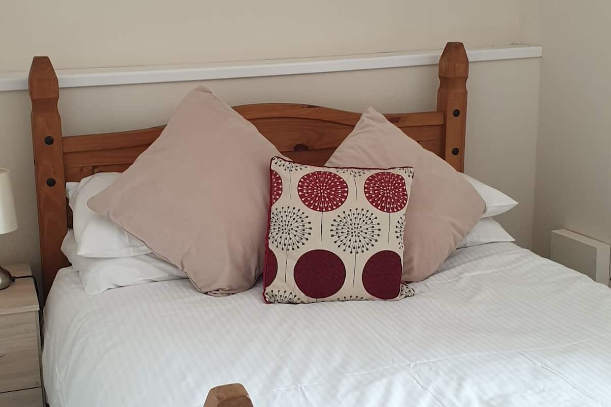 Una cama de madera con ropa de cama blanca y dos almohadas, una lisa y otra con estampado rojo y blanco.