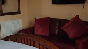 A hotel room with a bed, a wooden headboard, a brown leather sofa with red cushions, a mounted TV, and a radiator.