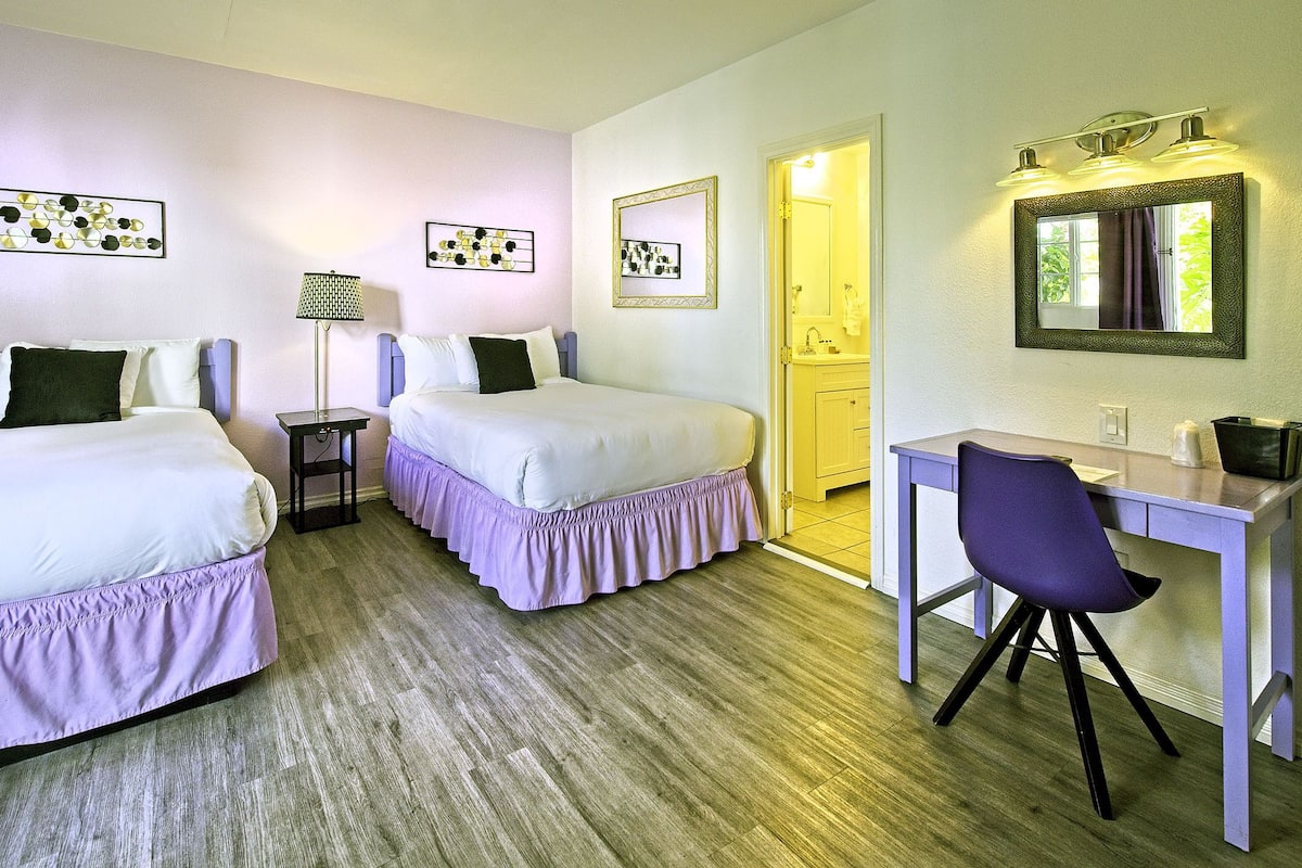 Une chambre d’hôtel avec deux lits, un bureau et une chaise. Les murs sont ornés de tableaux encadrés et un miroir est placé au-dessus du bureau.