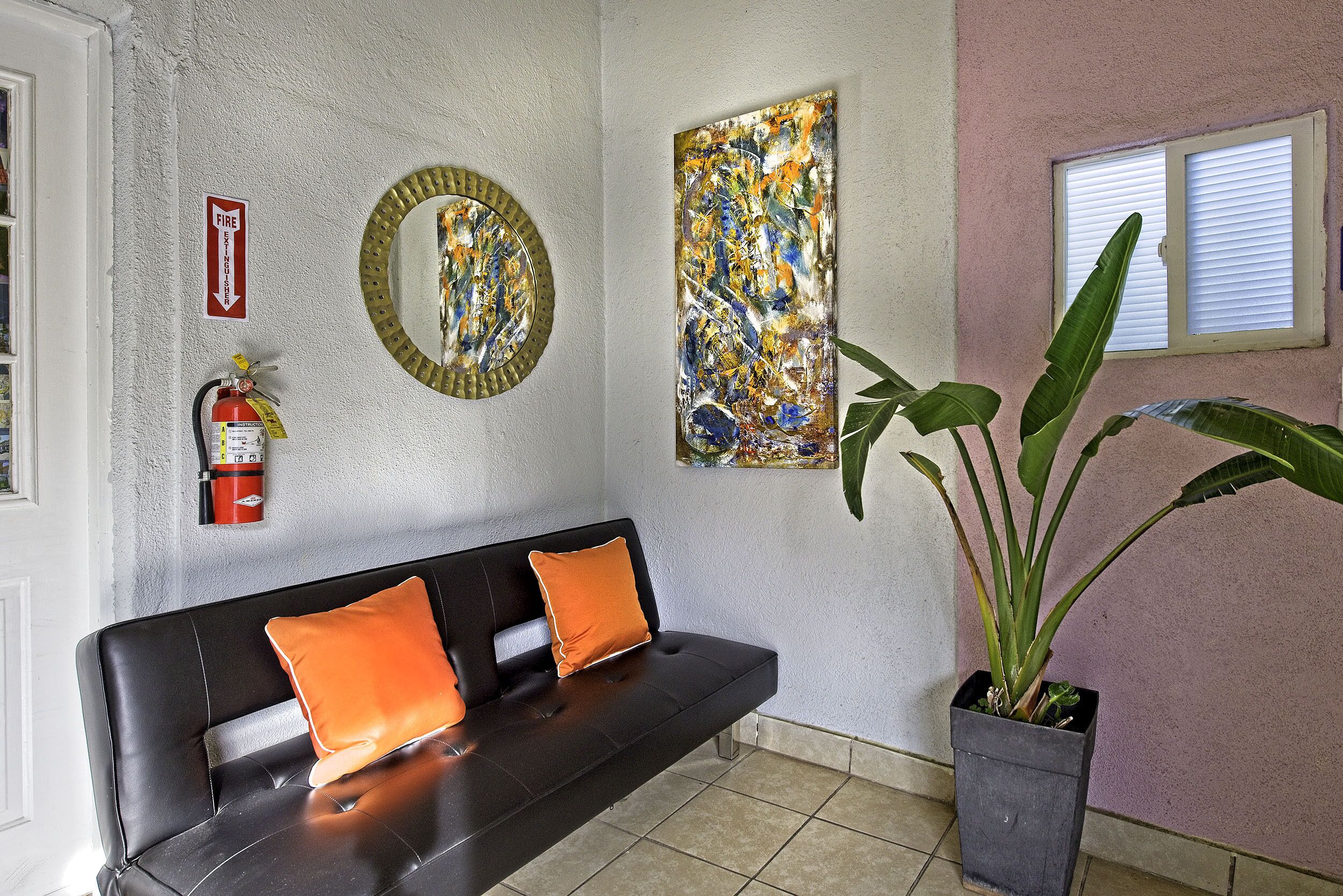 A black leather sofa with orange cushions, a fire extinguisher, a circular mirror, and a large abstract painting on the wall.