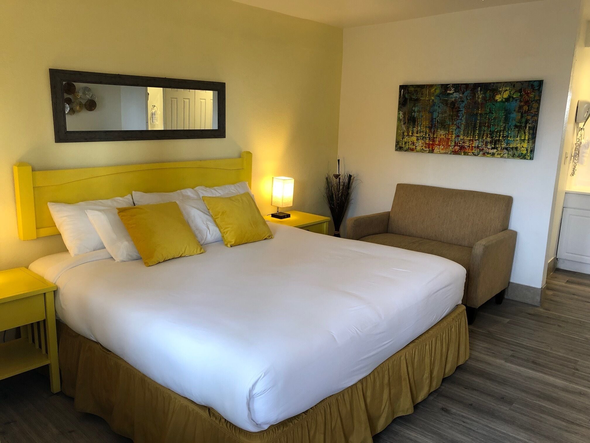 Ein Hotelzimmer mit einem großen Bett, einer gelben Kopfteil und einem braunen Sessel. An der Wand hängt ein Gemälde und auf dem Nachttisch steht eine Lampe.